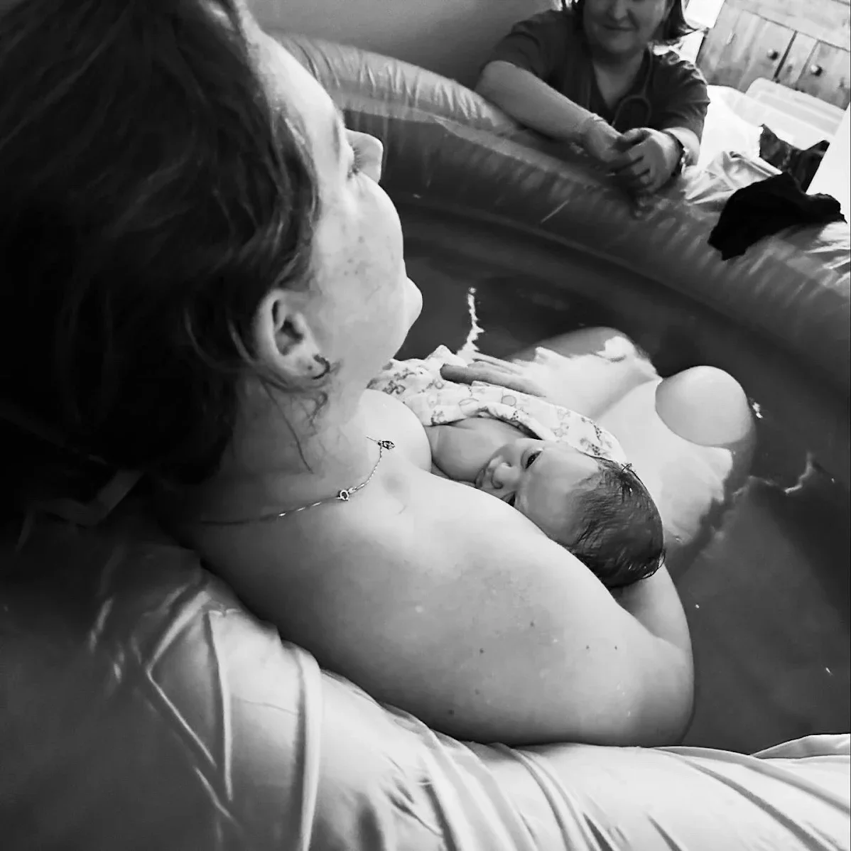 A woman holding a newborn baby in a small inflatable pool, with a woman sitting nearby on the other side of the pool, in a cozy room.
