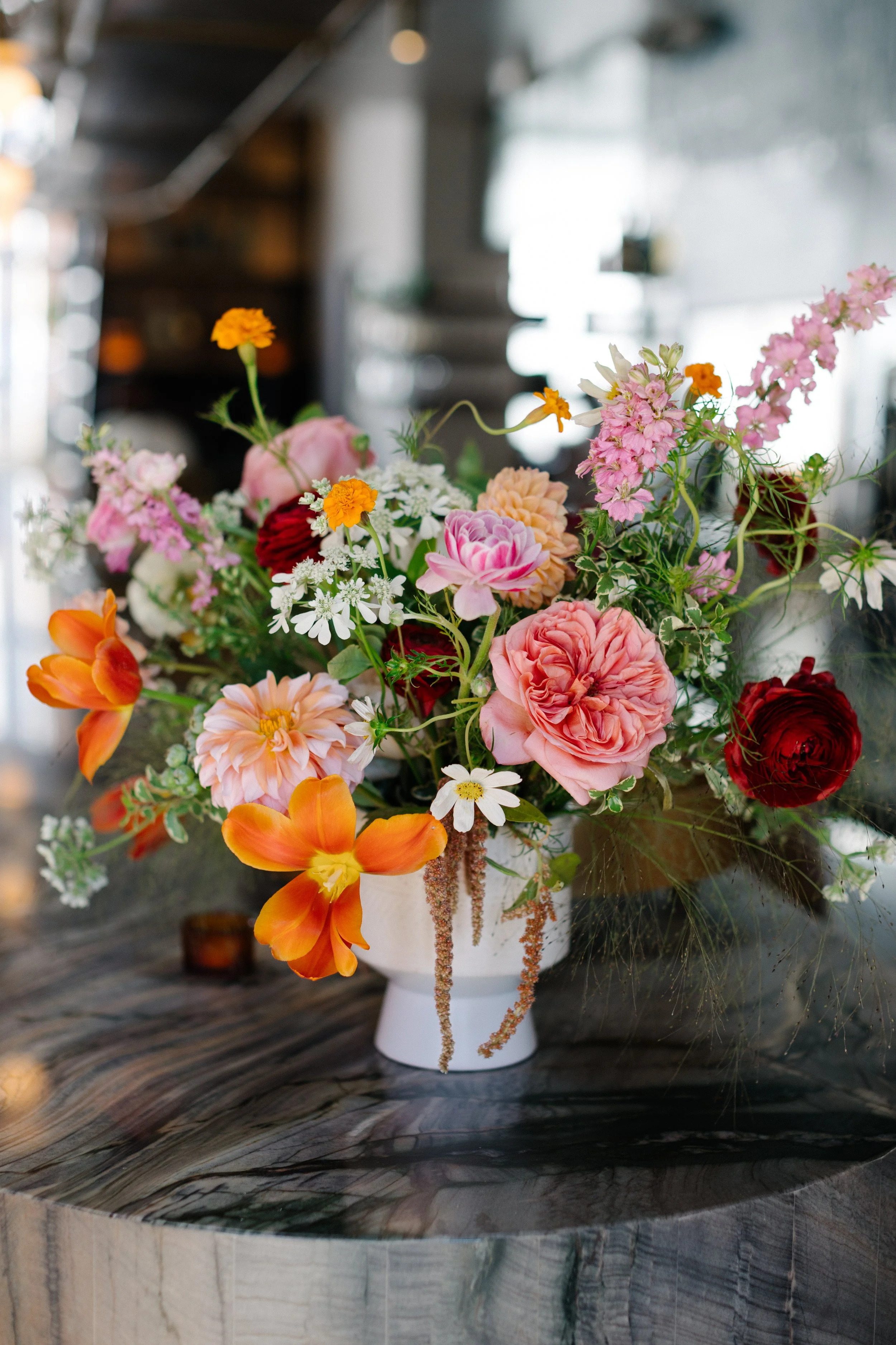 A colorful bouquet of various flowers in a white vase on a bar top, with a blurred indoor background.