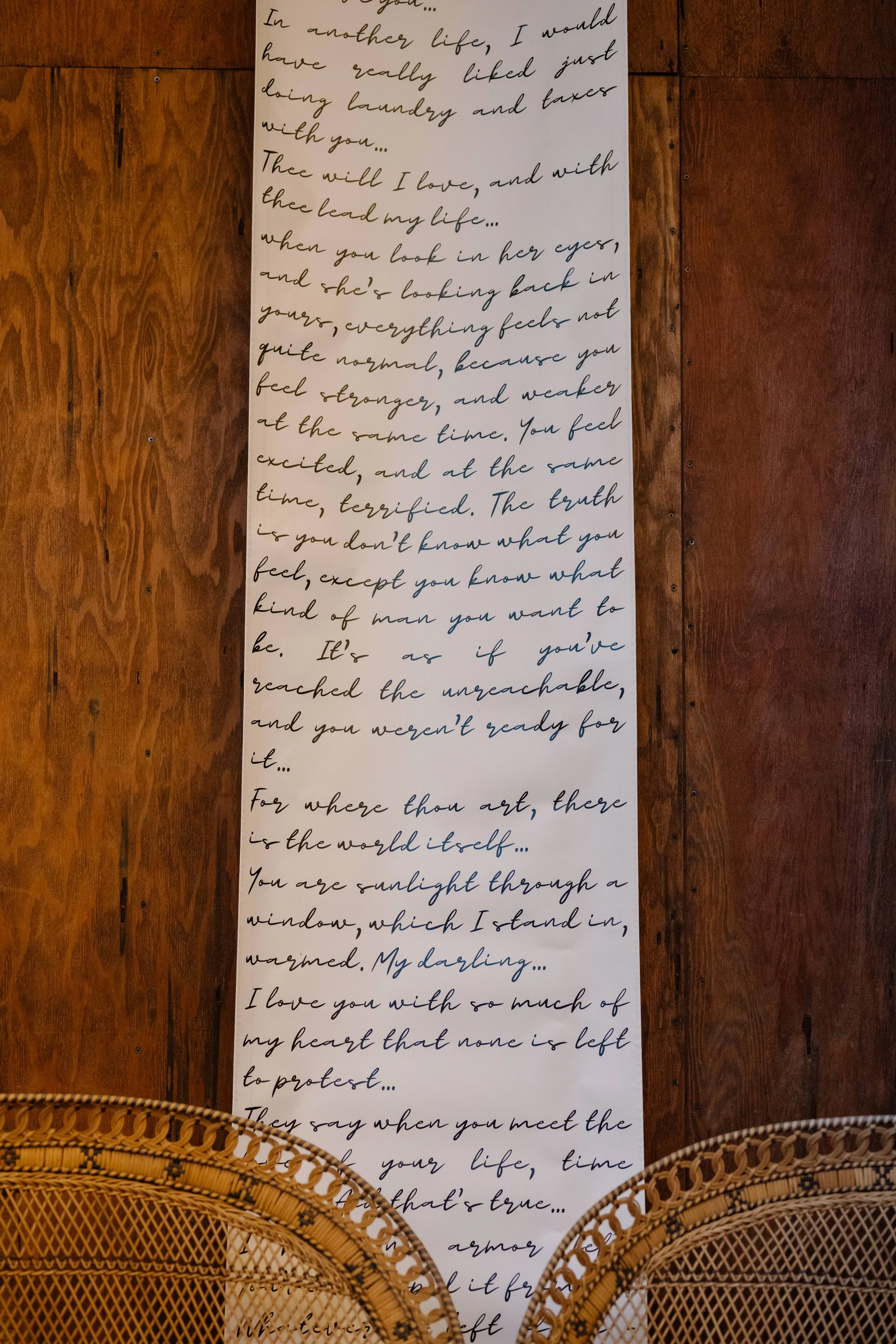 A long white ribbon or banner with handwritten cursive text in black ink, placed vertically on a wooden surface. The text appears to be a romantic or poetic message, and there are two woven rattan chairs at the bottom of the image.