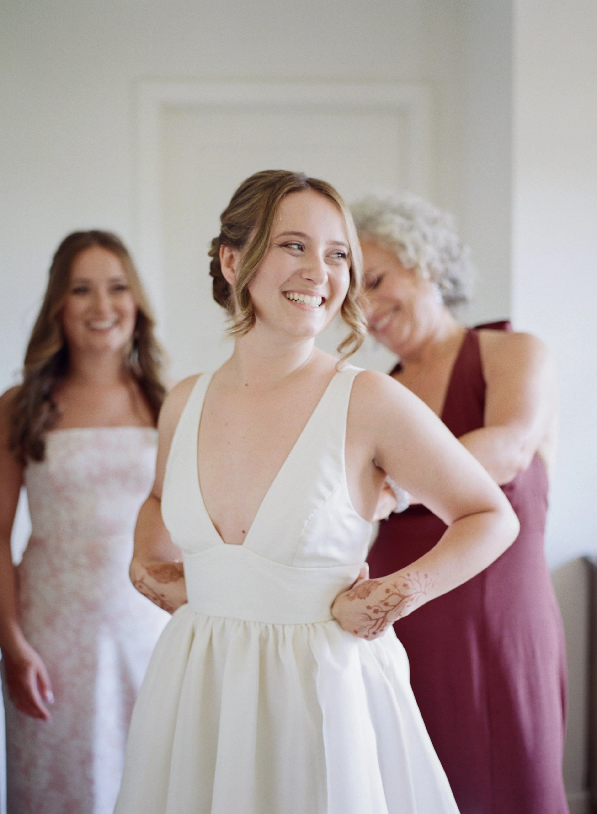A bride in a white wedding dress smiling with her hands on her hips, while two women zip the dress up behind her, one in a floral dress and one in a burgundy dress, in a bright room.