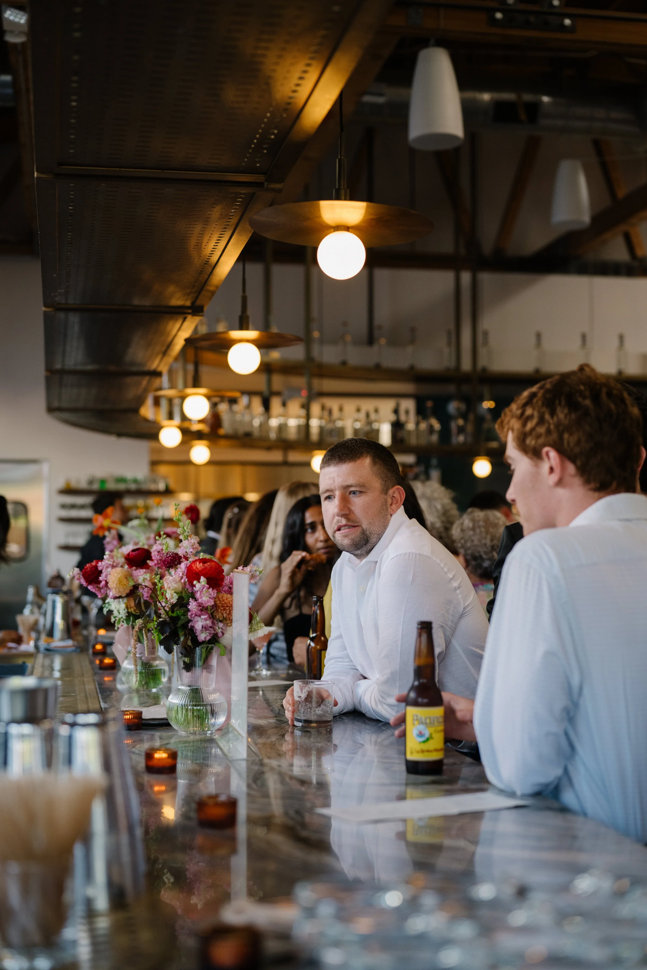 People sitting at a bar in a restaurant, engaging in conversation, with floral arrangements and bottles on the counter, illuminated by warm pendant lighting.