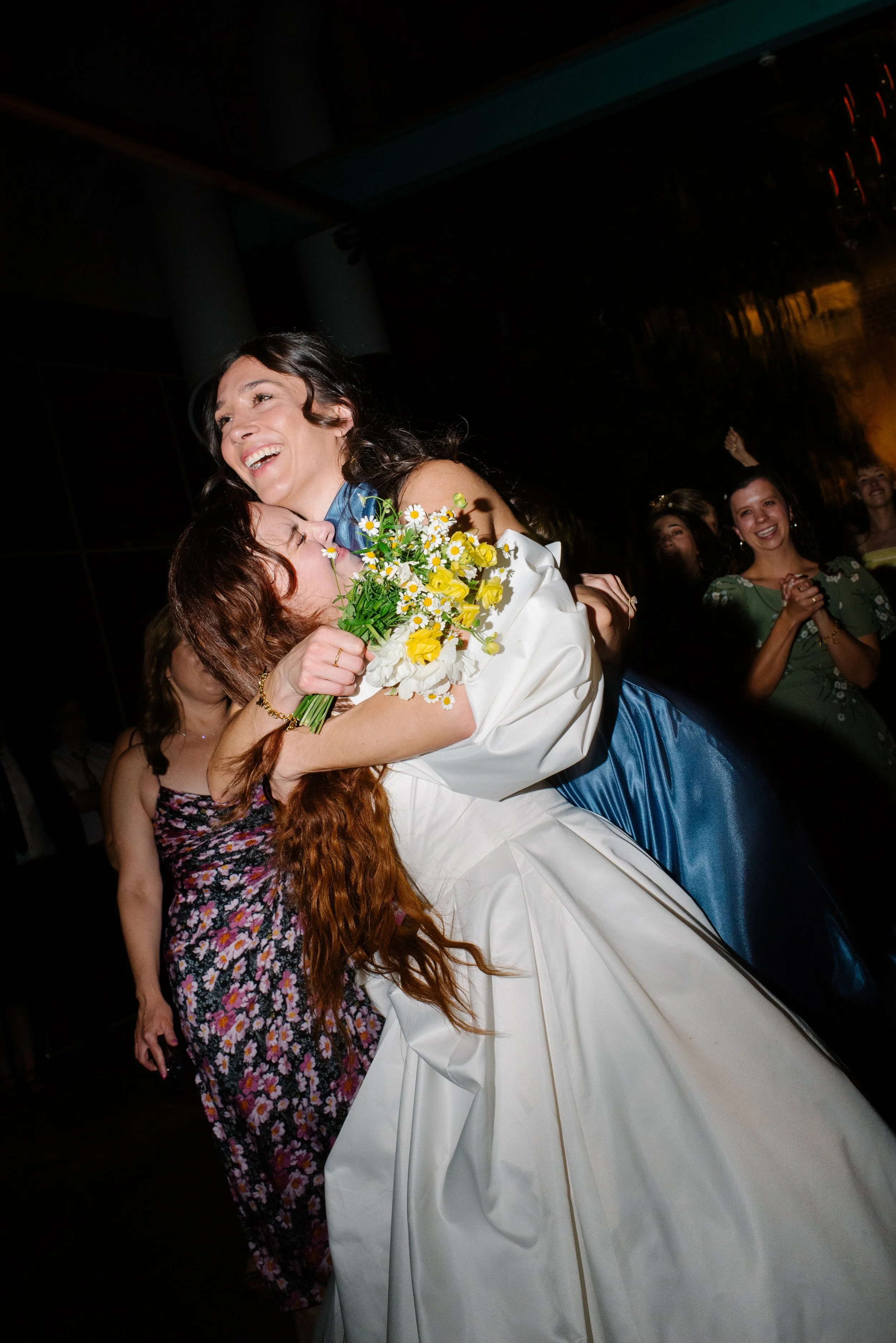 A bride hugging and picking up a woman in a blue dress, who is holding a bouquet of yellow and white flowers, others smiling and clapping in the background.