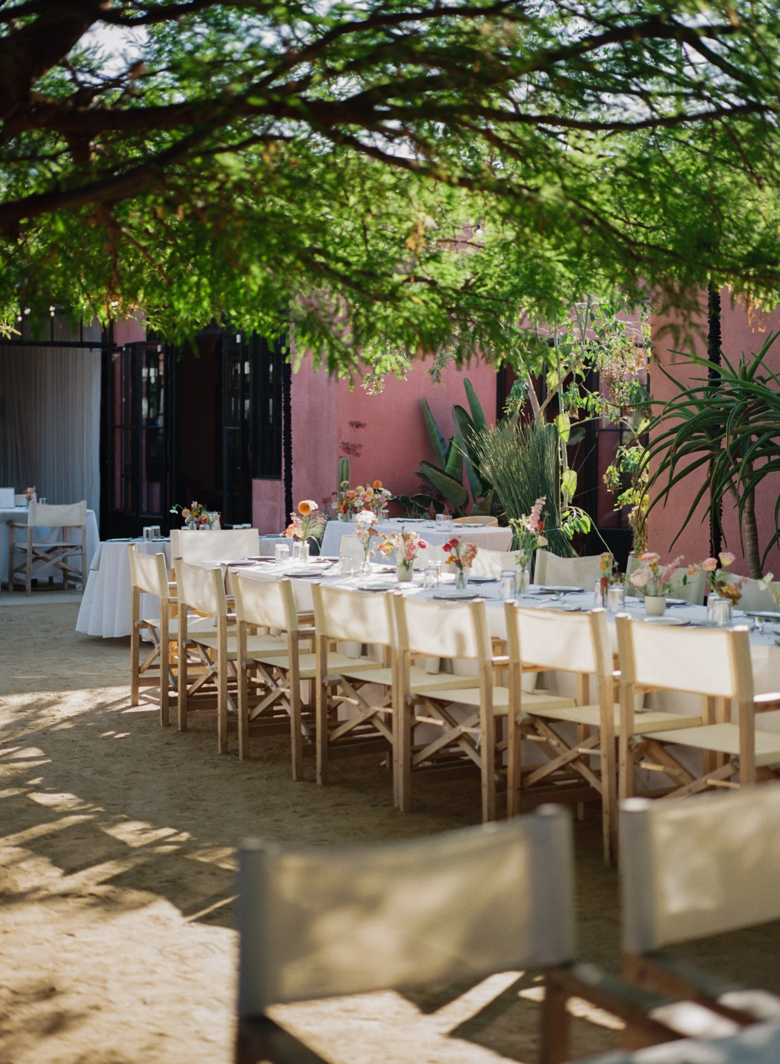 Outdoor dining area with long table set with flower arrangements, surrounded by wooden chairs, shade from trees, pink wall and plants in the background.