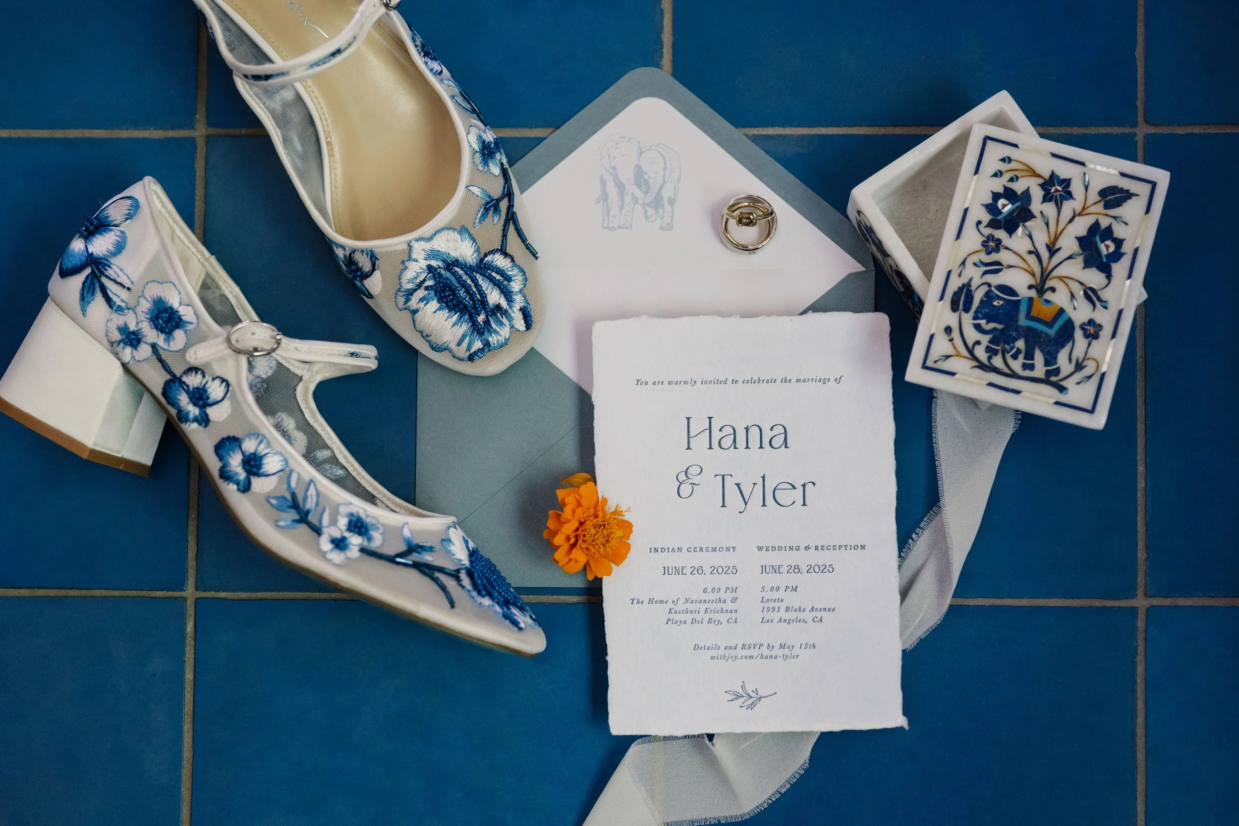 Wedding invitation surrounded by white and blue embroidered shoes, a ring, a flower, and decorative cards on a blue tiled floor.