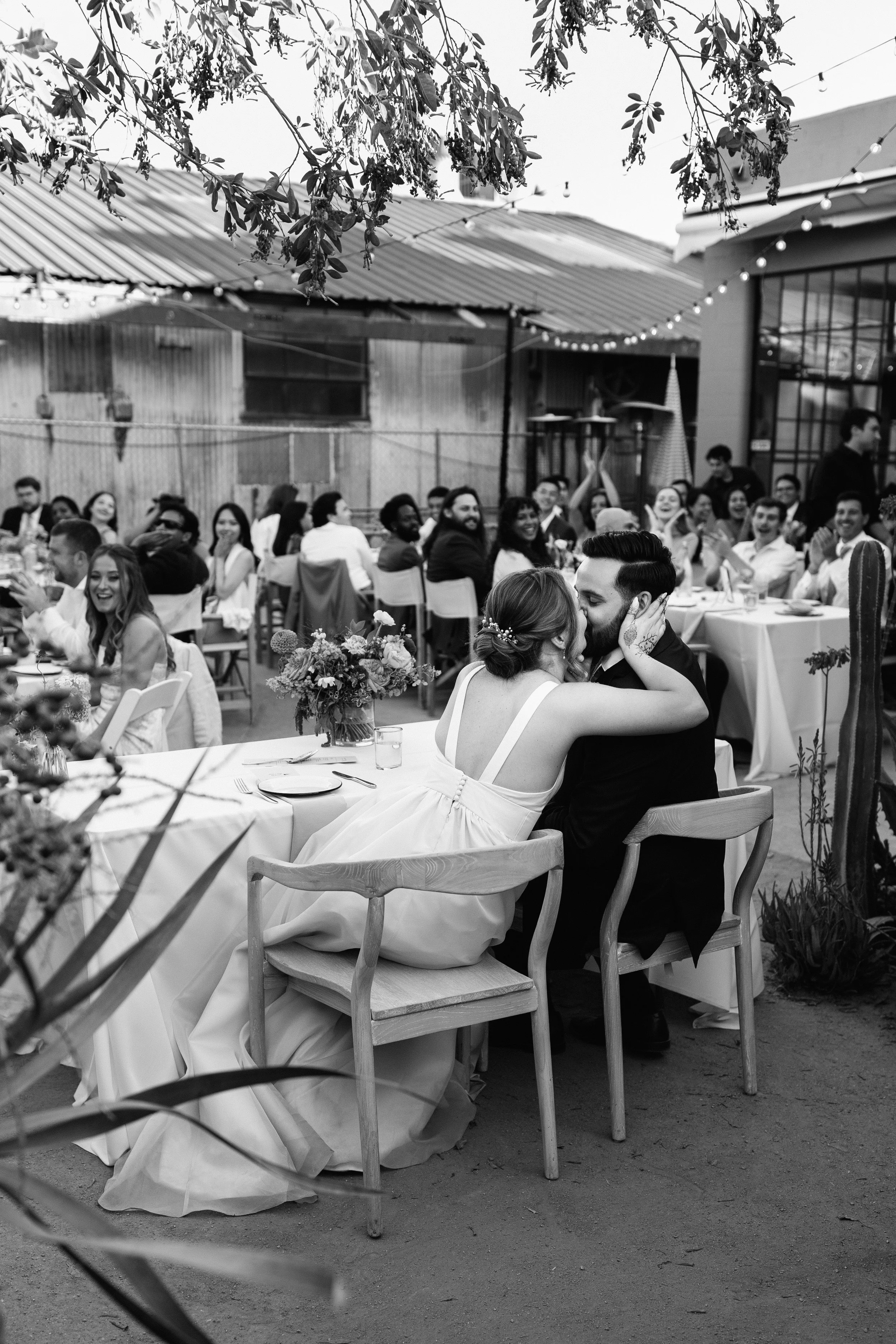 A black and white photo of a wedding reception with a bride and groom sharing a kiss at their table, surrounded by guests who are clapping and smiling.