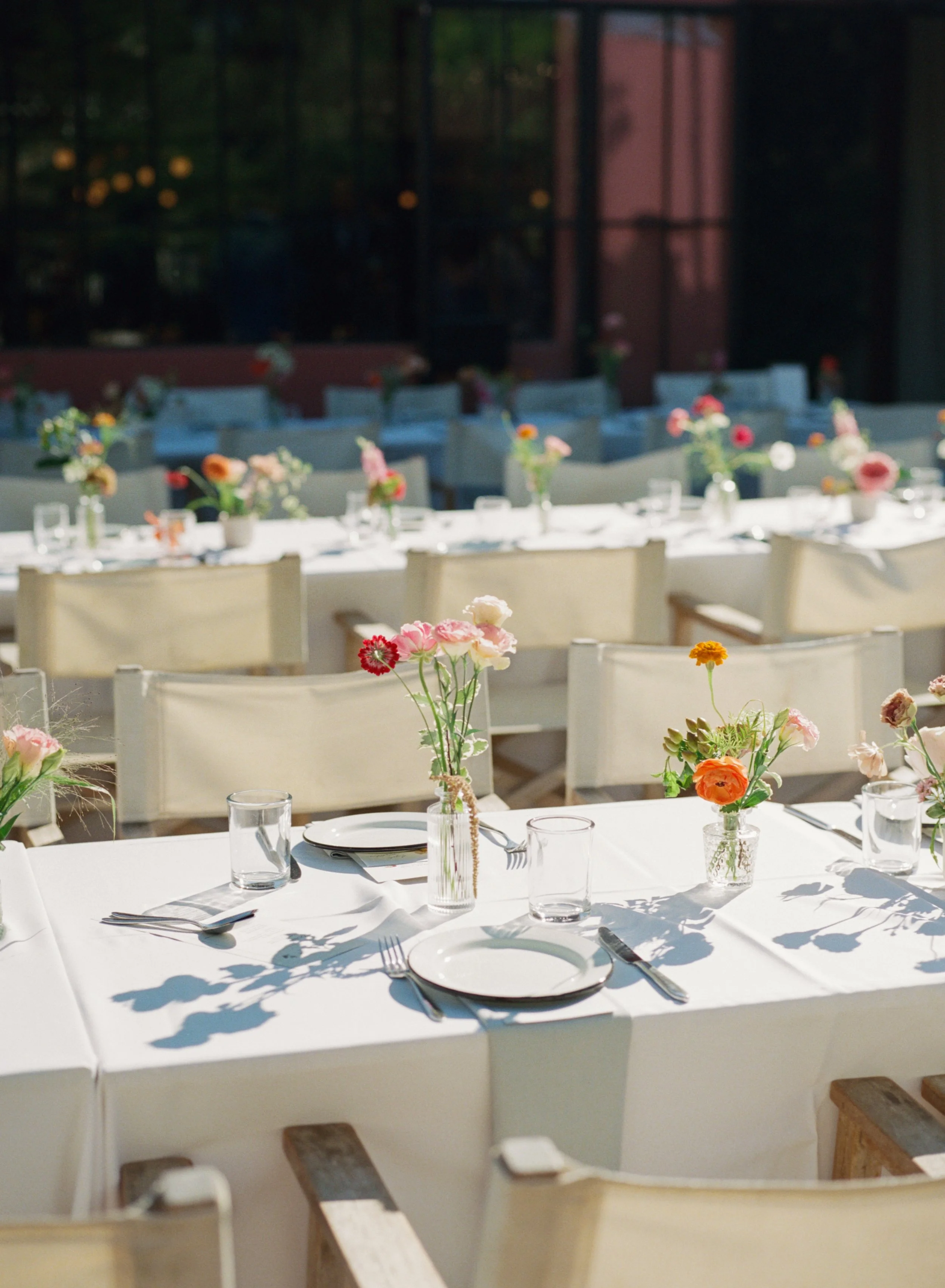 Elegant outdoor dining setup with white tablecloths, plates, glassware, silverware, and centerpieces of colorful flowers in vases, illuminated by natural sunlight.