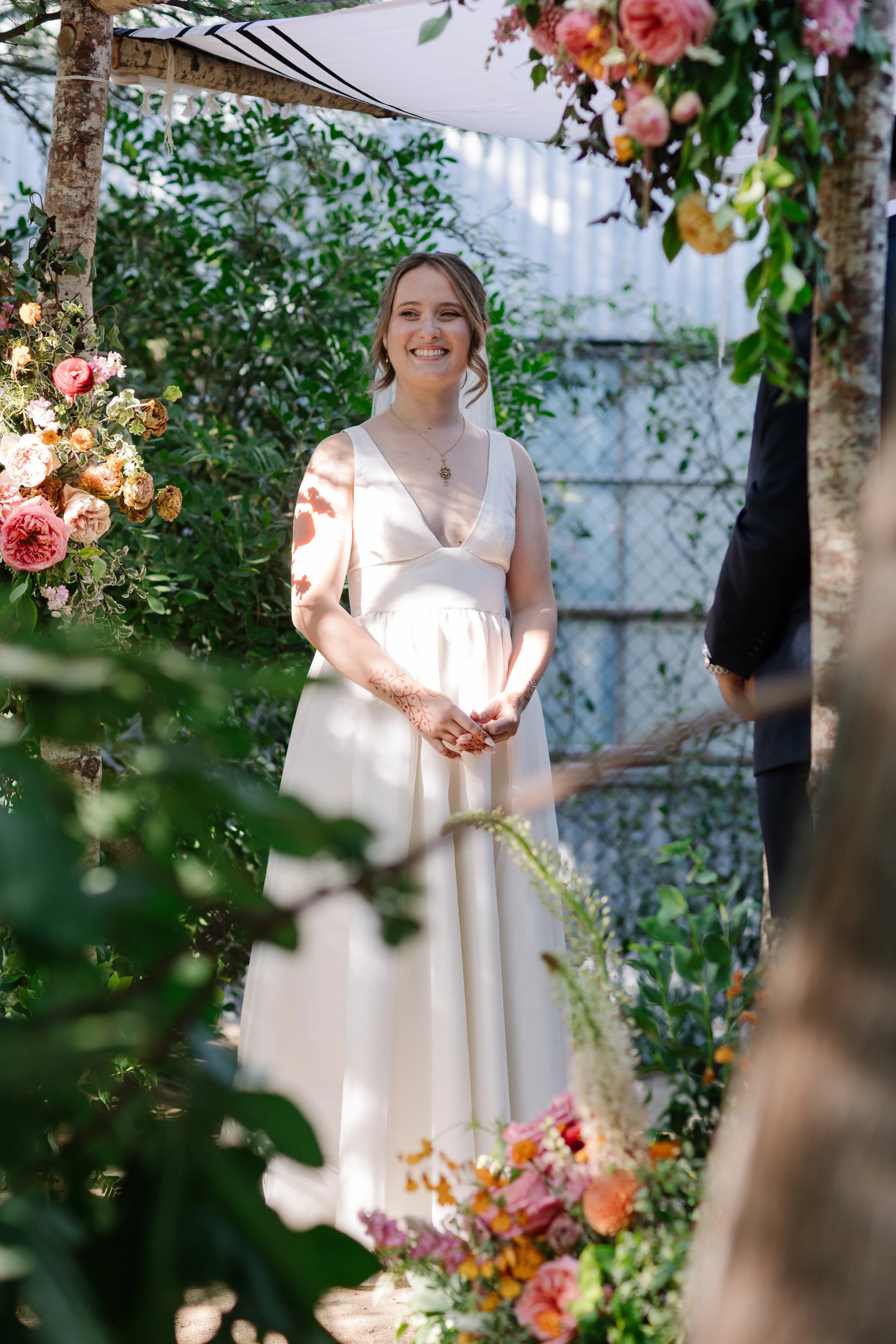 A woman in a white wedding dress smiling during a wedding ceremony outdoors under a Jewish chuppah surrounded by greenery and decorated with colorful flowers.
