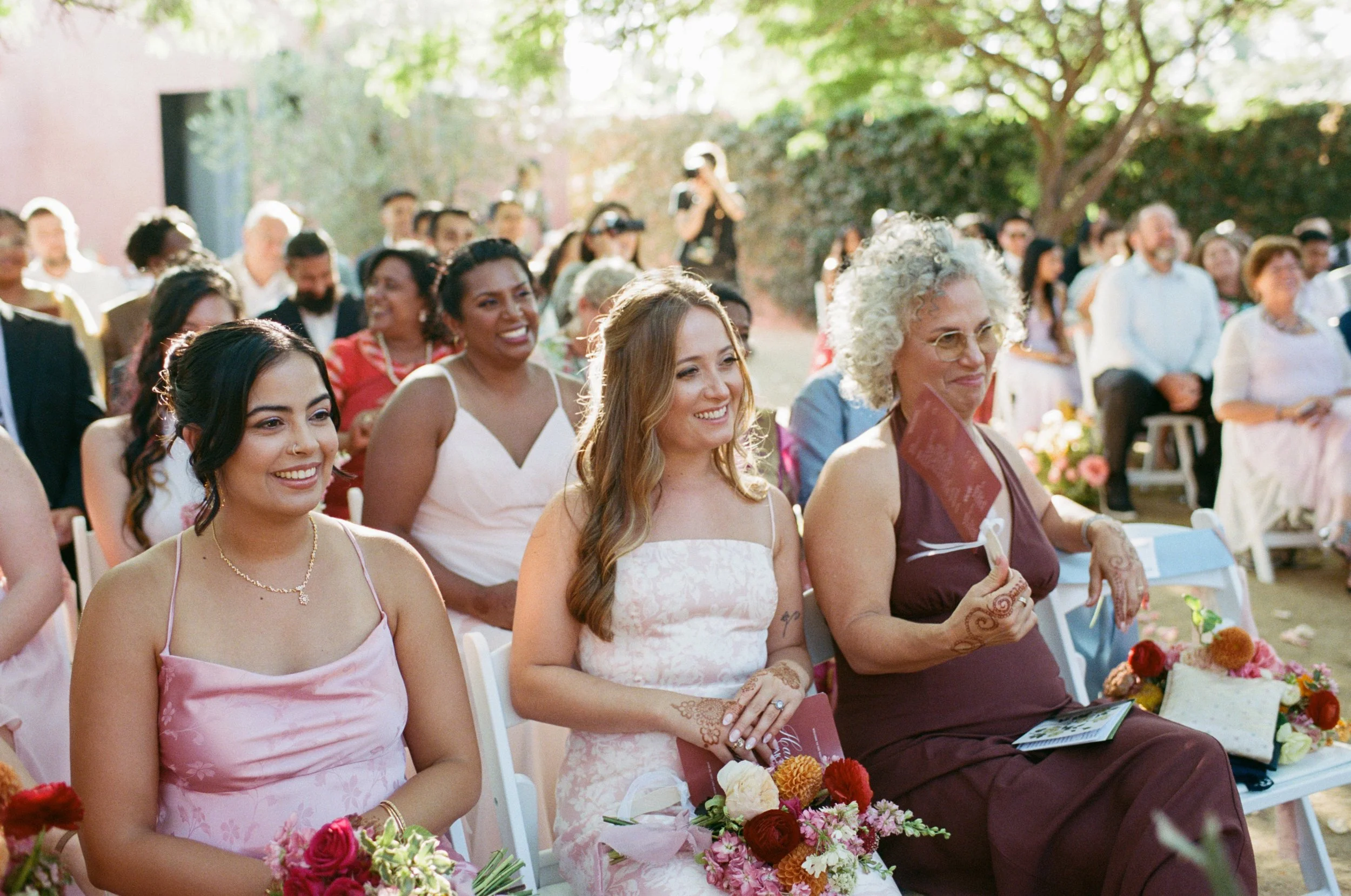 An outdoor wedding ceremony with smiling guests seated on white chairs, holding bouquets of colorful flowers, under a tree with green leaves, during the daytime.