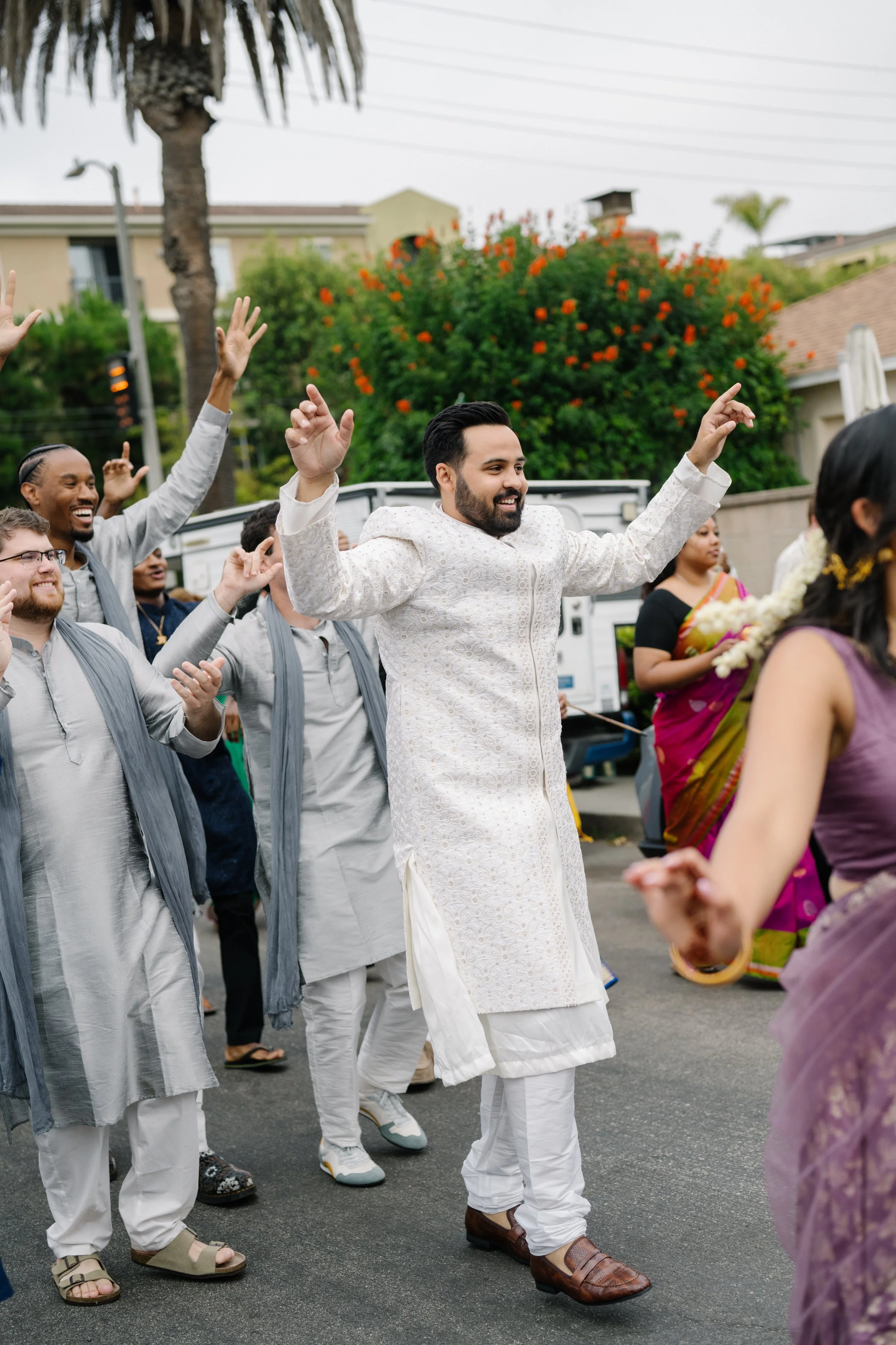 A group of people celebrating and dancing outdoors during a traditional Indian wedding procession or baraat, with men dressed in kurta and dhoti, and women in sarees.