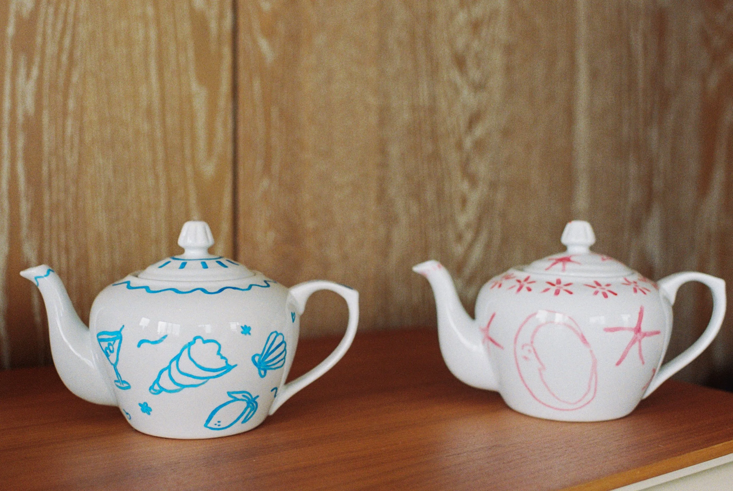 Two white ceramic teapots with hand-drawn designs, one with blue decorations and the other with pink, placed on a wooden surface against a wooden wall background.