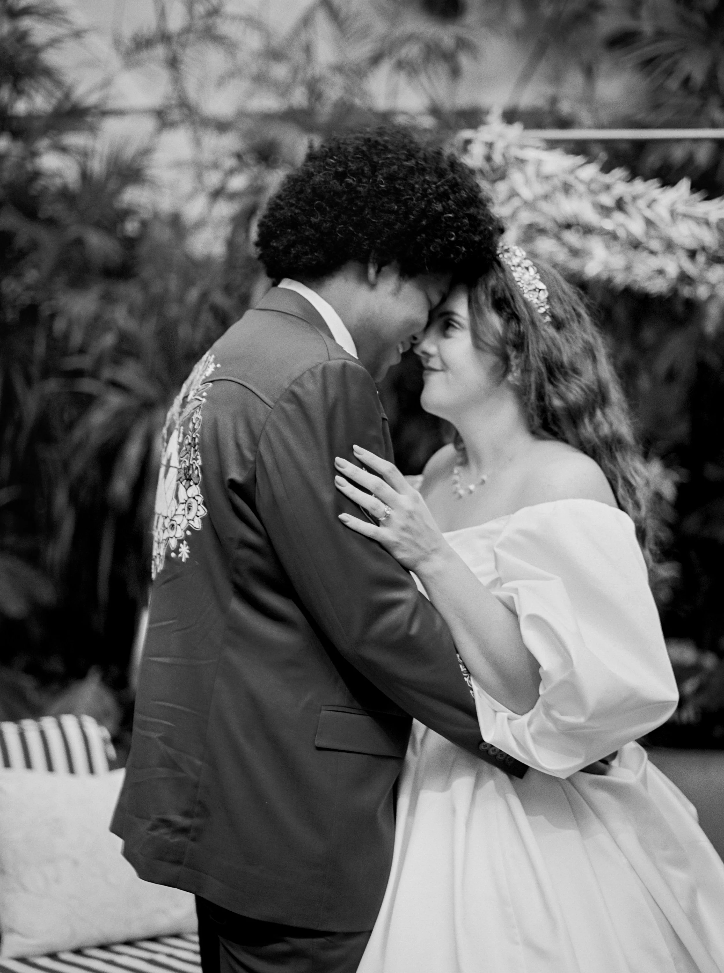 A black and white photo of a couple touching foreheads and smiling, dressed in wedding attire, in a lush, garden setting.