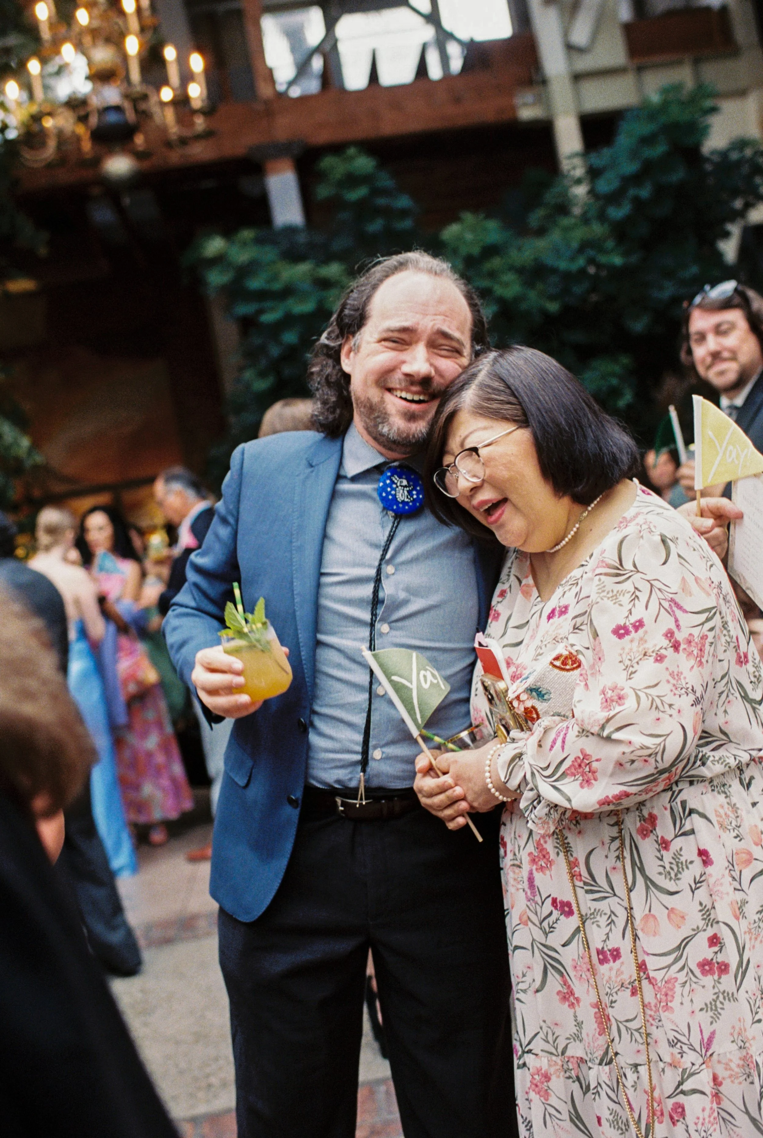 Two people, a man in a blue suit and a woman in a floral dress, smiling and enjoying a wedding reception, holding small flags and drinks, with other guests in the background.