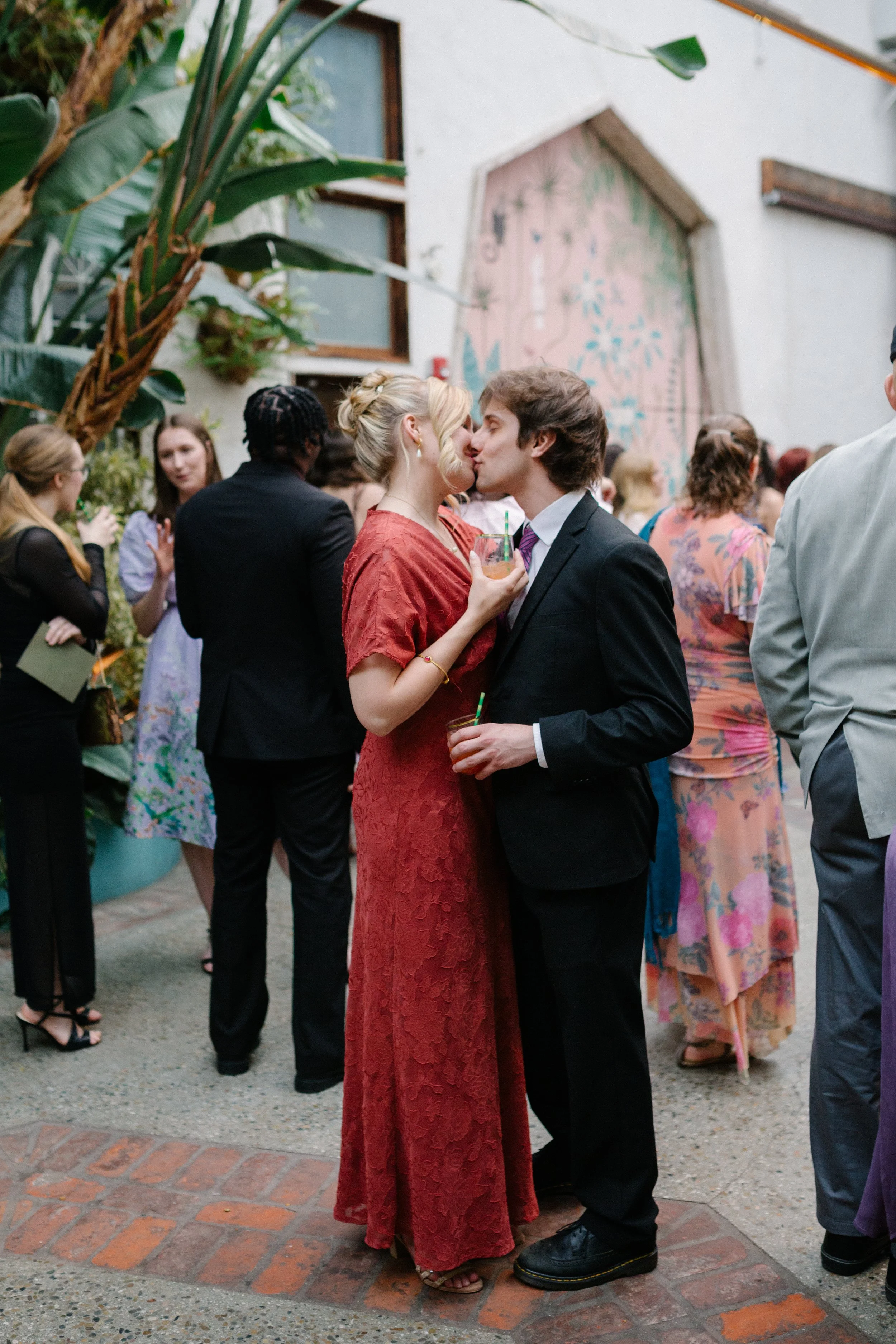 A man and woman kissing at a wedding cocktail hour, with other attendees in the background.