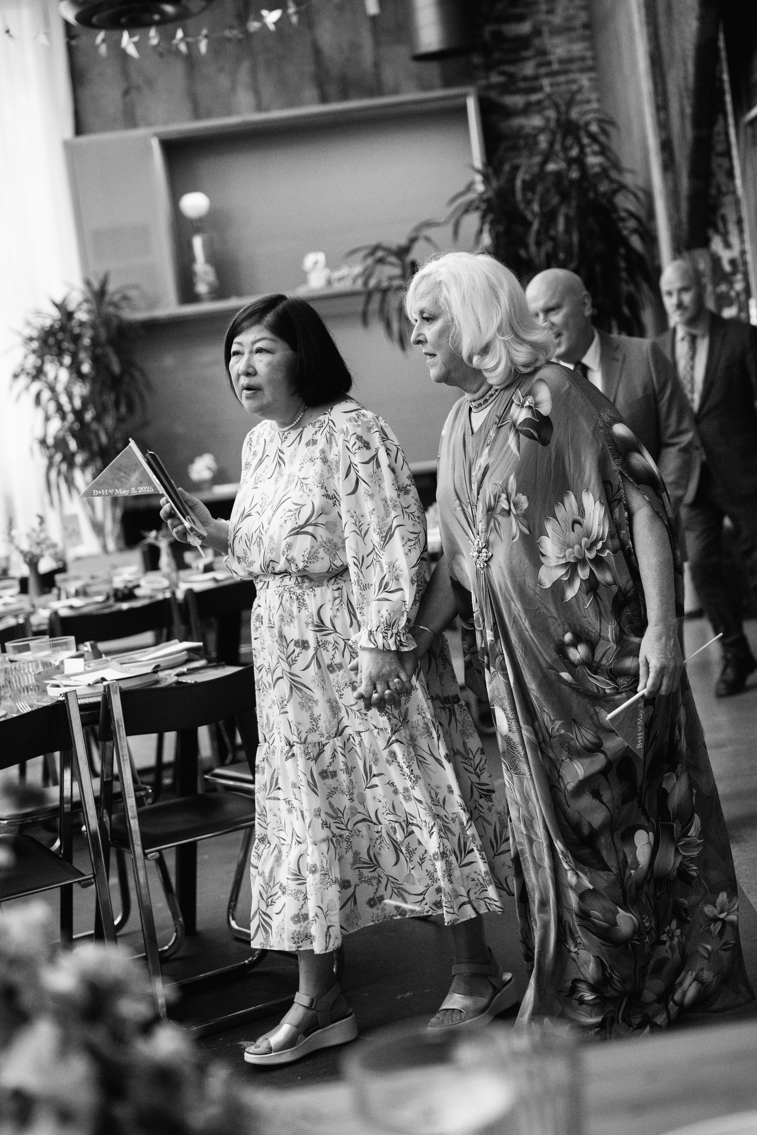 Two women holding hands and walking together at a wedding, with several other people in the background, in a room decorated with plants and table settings.