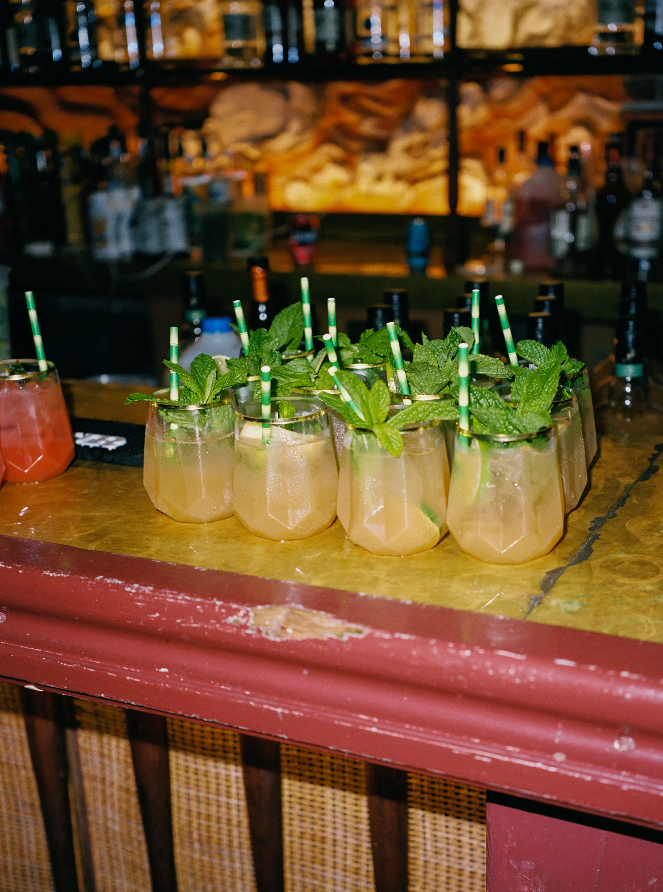 A group of mint garnished cocktails on a wooden bar counter with a blurred bar scene in the background.