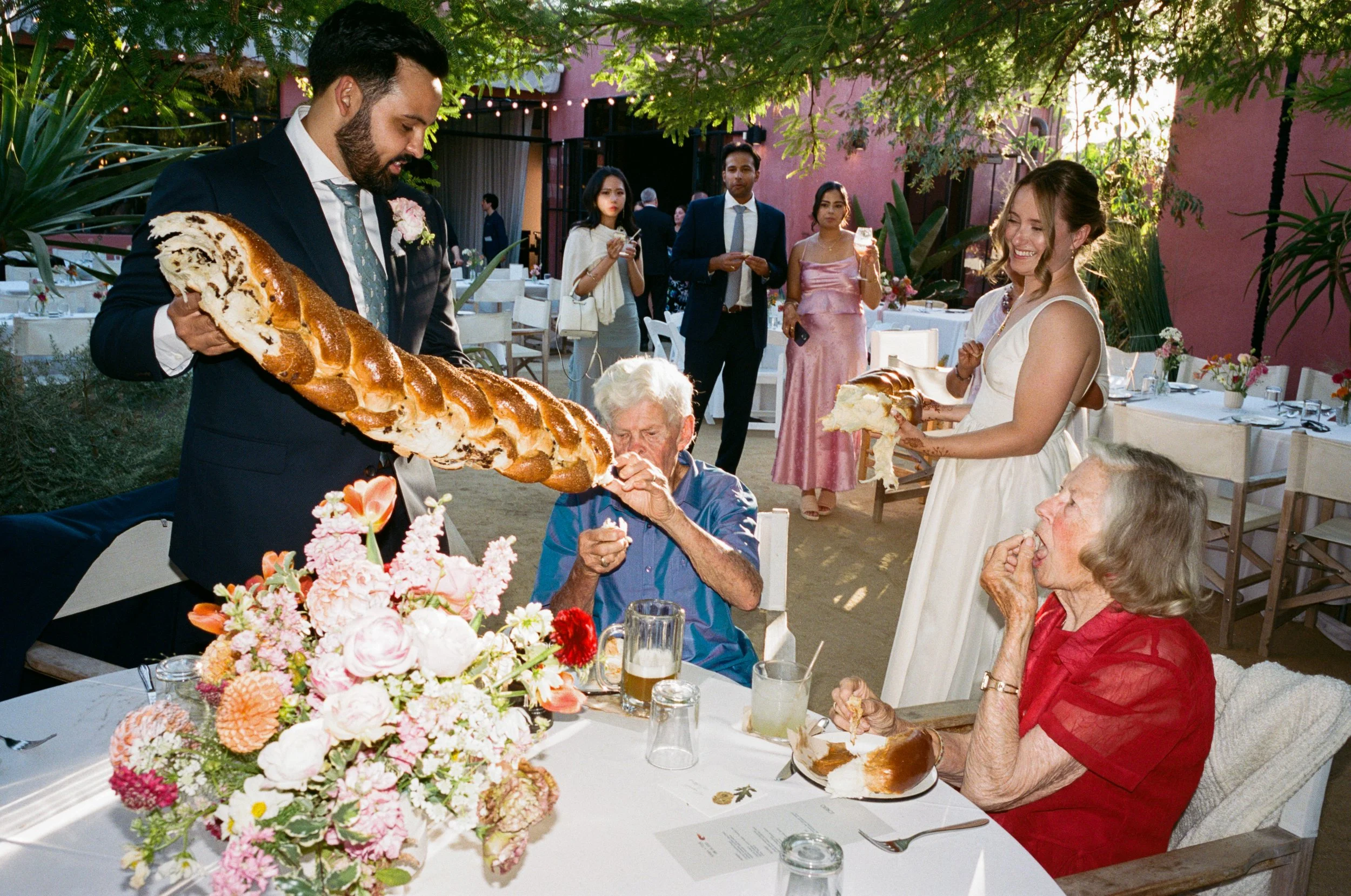 A man with a beard in a suit is holding a large braided loaf of challah bread at an outdoor Jewish wedding. Two elderly people are seated at a table, eating slices of bread. .