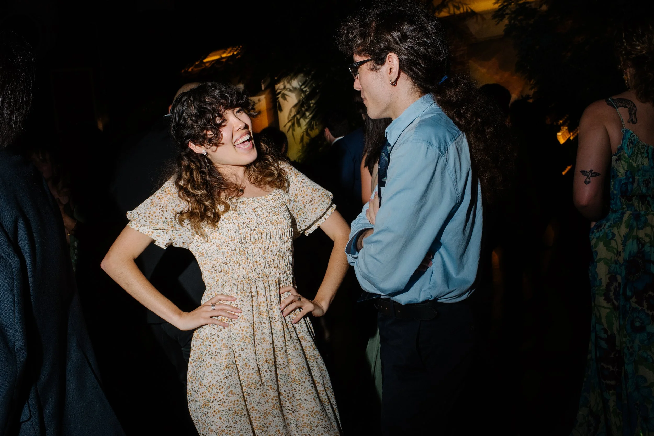 A woman with curly brown hair smiling and talking with a man with glasses at a wedding.