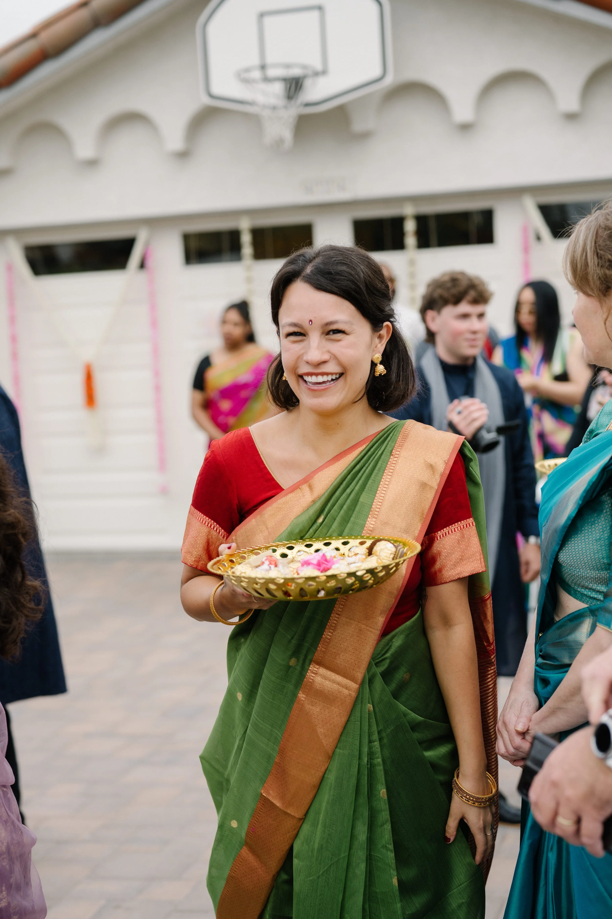A woman dressed in a traditional green and orange saree smiling and holding a golden tray with flowers. She is at an outdoor baraat with other guests in the background.