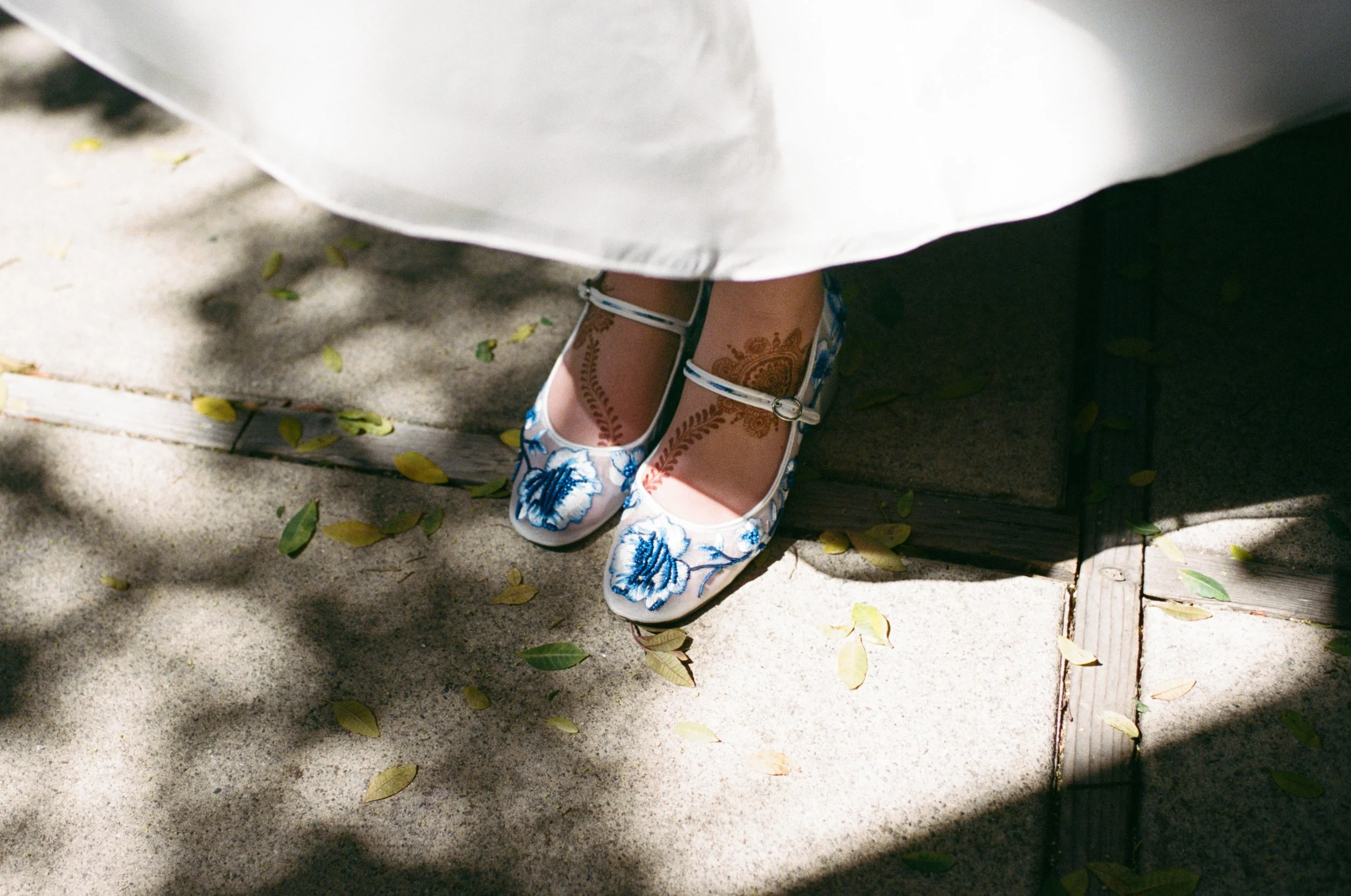 Close-up of a person's feet wearing traditional embroidered shoes, standing on a concrete sidewalk next to a wooden platform, with a white dress and scattered leaves and shadows around.