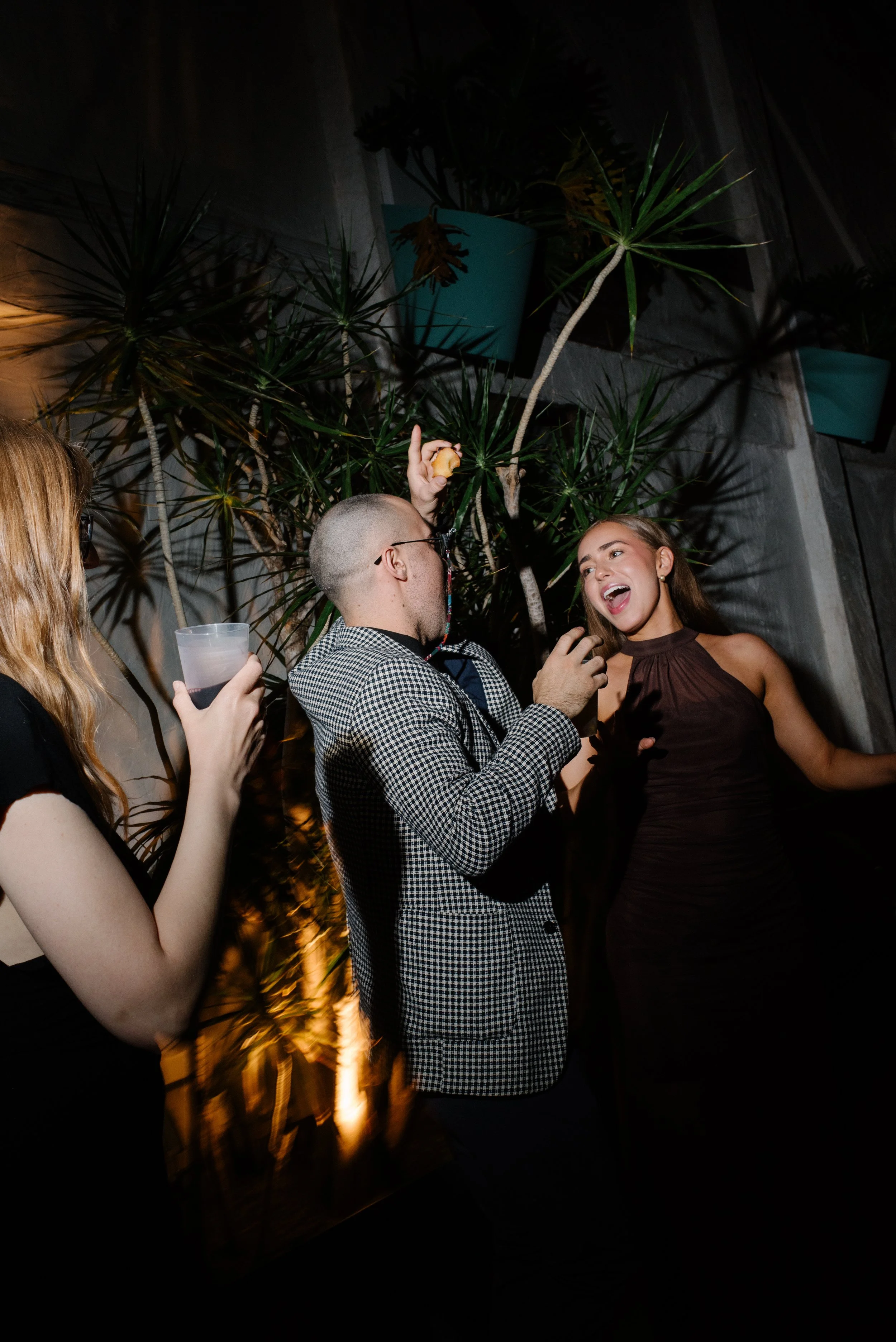 People dancing at a wedding, two women and a man, engaging in conversation and laughter, with tropical plants in the background.