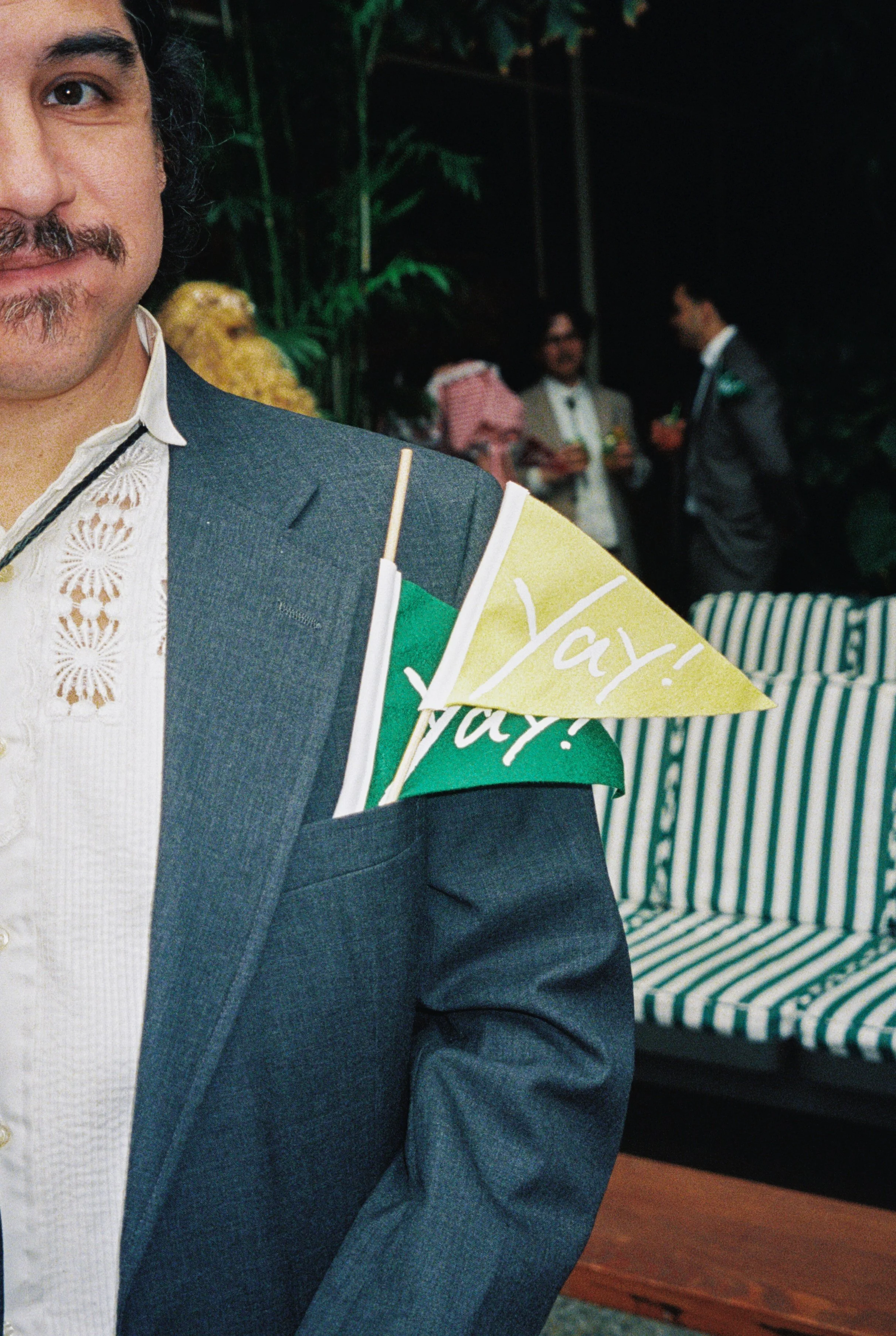 Close-up of a man in a gray suit at a wedding, with colorful flags in his lapel that say 'Yay!'. In the background, other people are socializing, and there is a striped outdoor sofa.