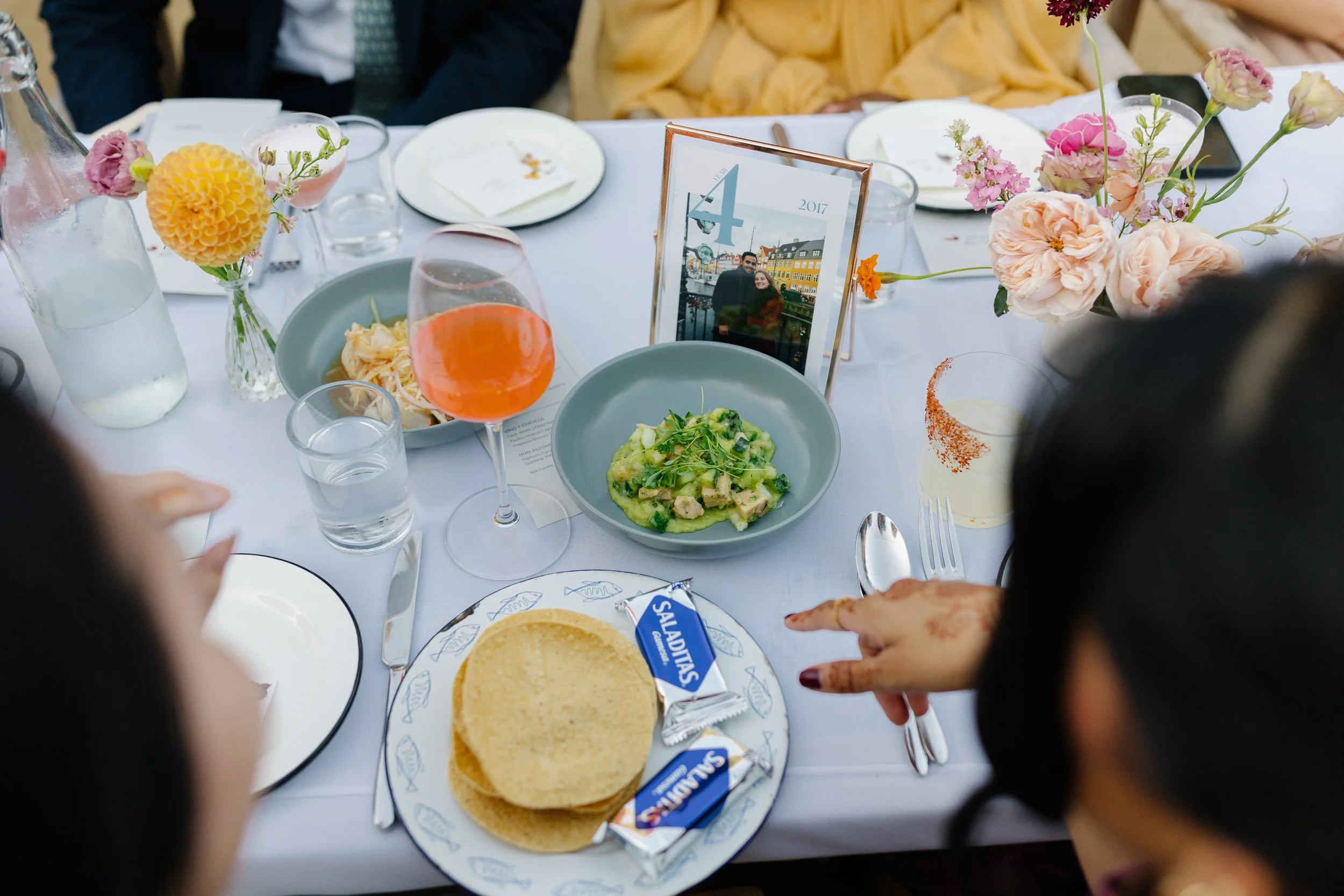 A dining table with plates of food, a glass of wine, salads, ceviche, tostadas, and floral centerpieces, with people seated around.