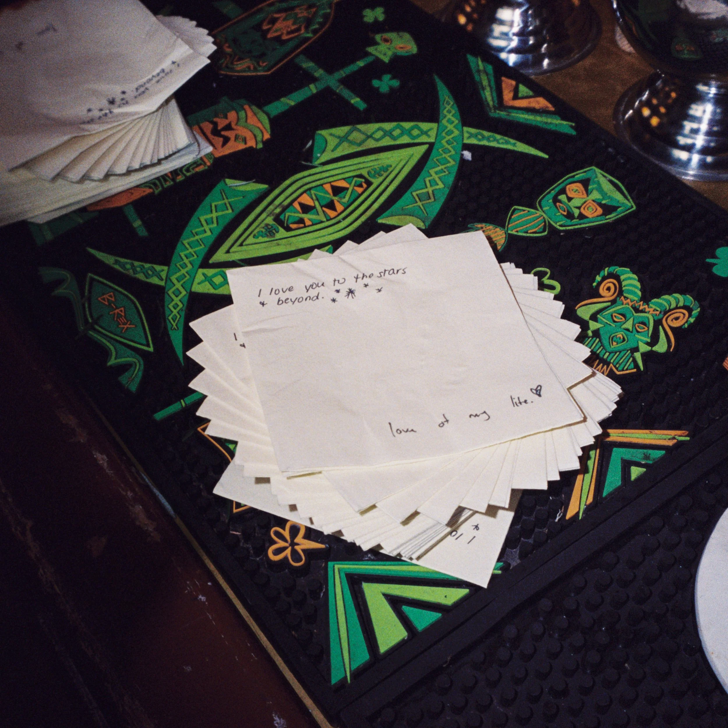 Custom napkins featuring handwritten text placed on a colorful, tribal-themed bar mat. The note says, 'I love you to the stars & beyond. Love of my life.' The bar mat has green, orange, and black patterns, including a face with horns.