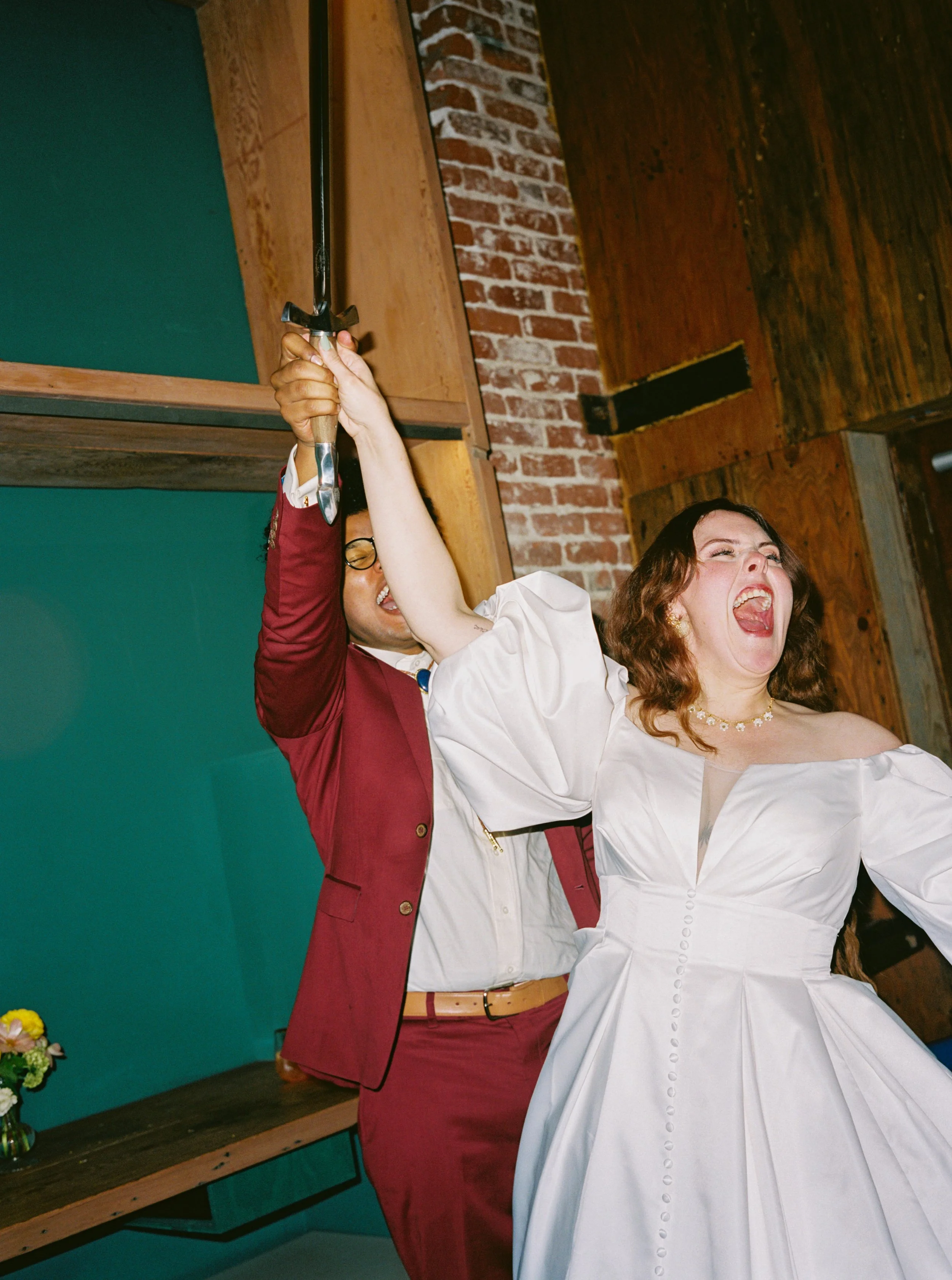 A woman in a white dress and a man in a red suit holding a sword together, celebrating joyfully after cutting their wedding cake.