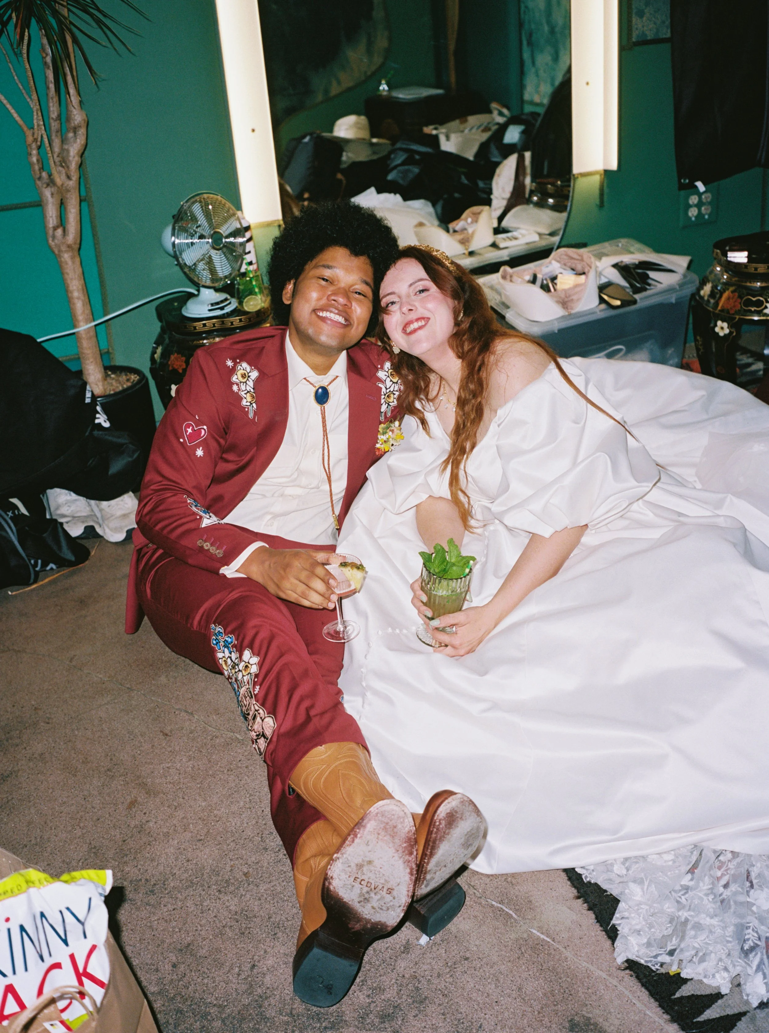 A joyful couple, with the man dressed in a embroidered red suit and cowboy boots, and the woman in a white wedding dress, sitting on the floor in a dressing room, smiling and holding drinks.