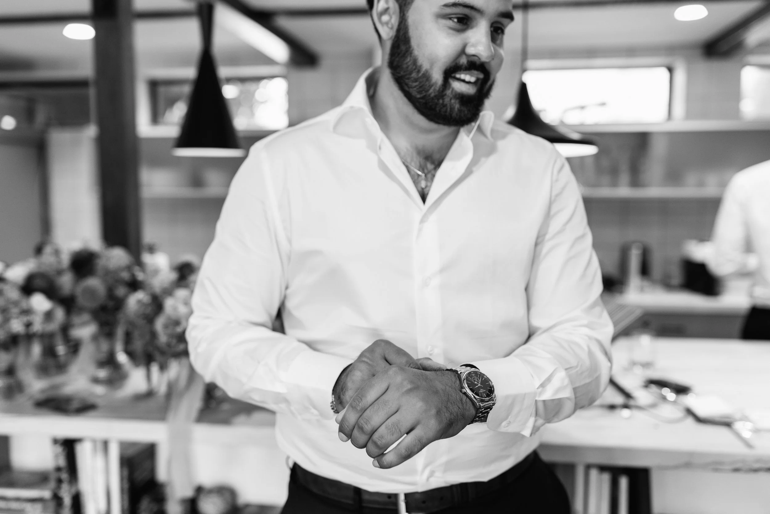 A man with a beard in a white dress shirt, standing in a room with natural light, with flowers and tables in the background.
