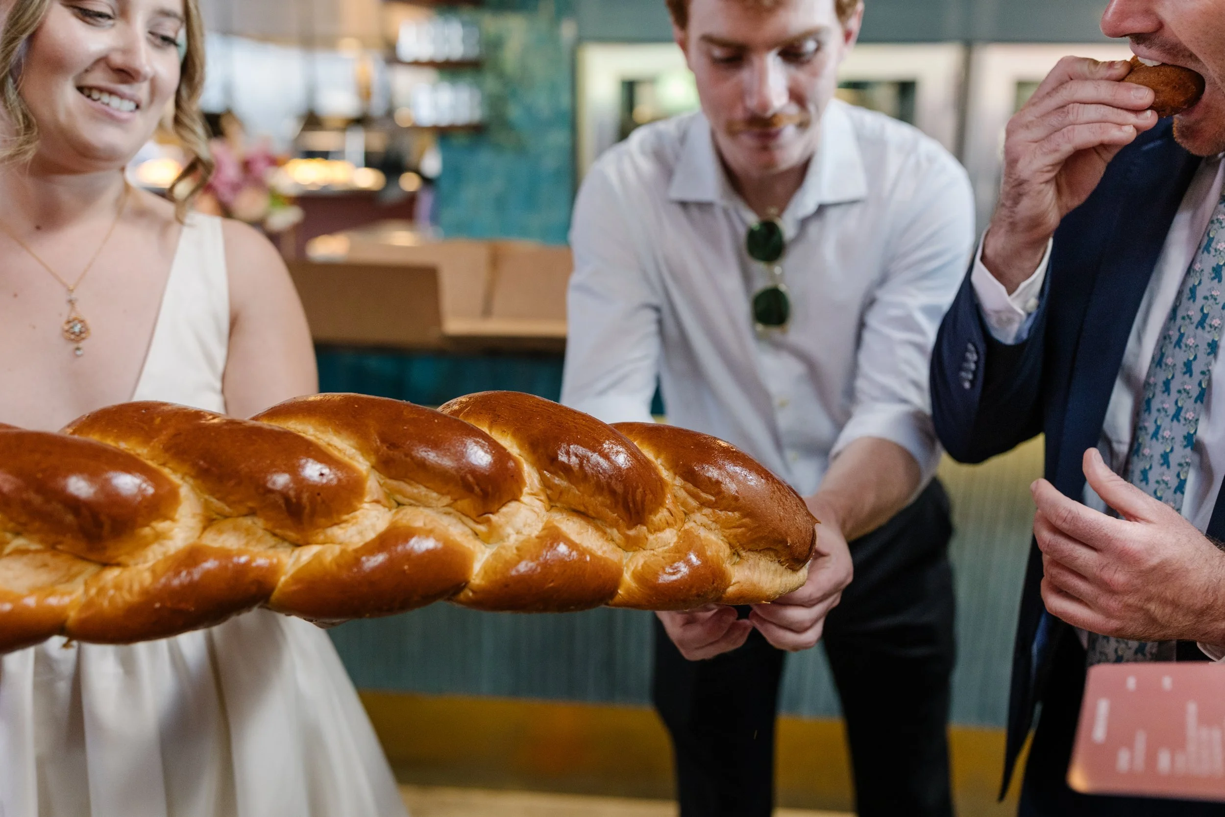 Three people, including a woman in a white dress, sharing a large braided loaf of challah bread.