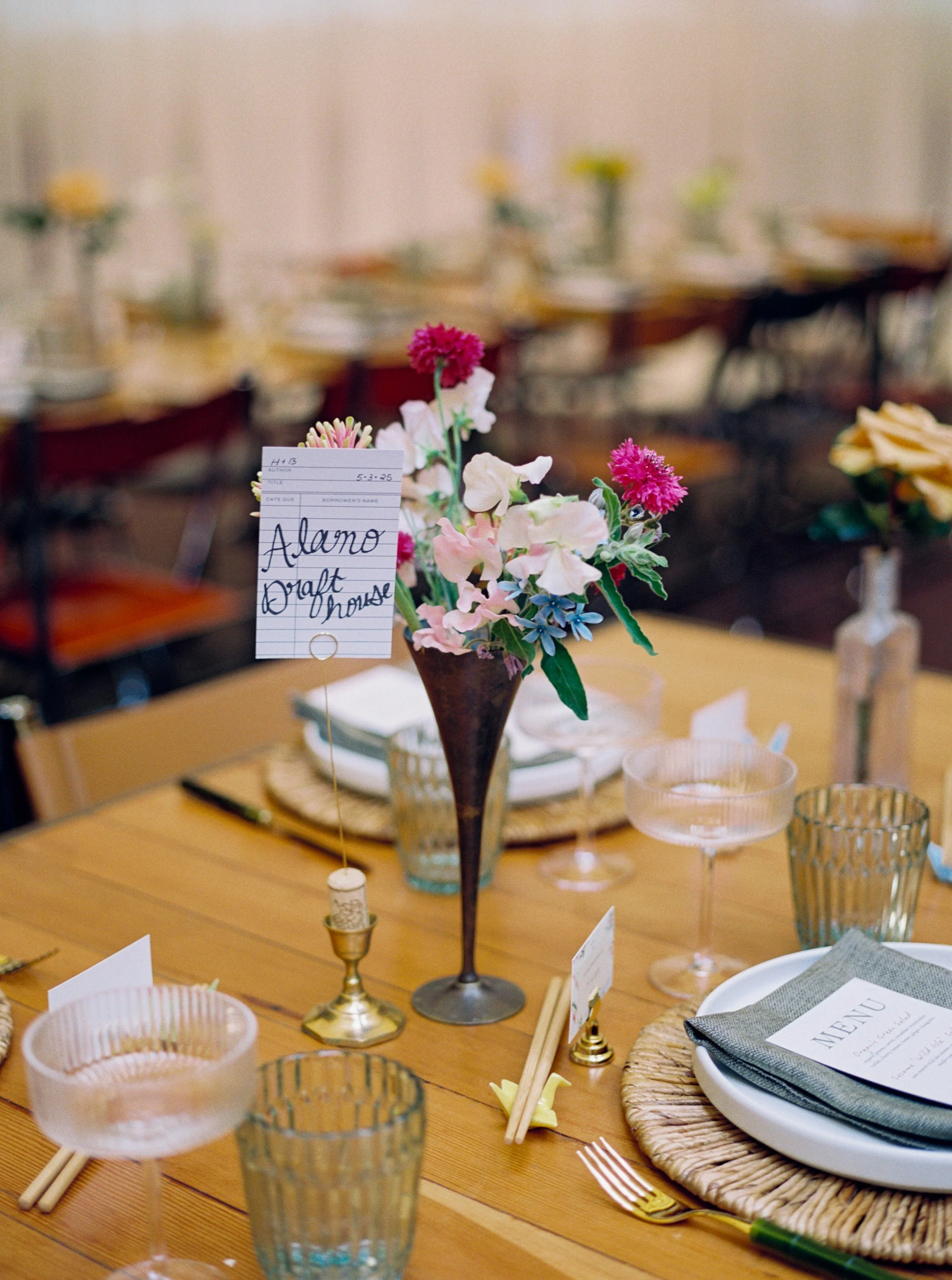 A decorated table with a pink flower arrangement, vintage glasses, plates, and cutlery, with a small table sign and a menu card, in a rustic setting.