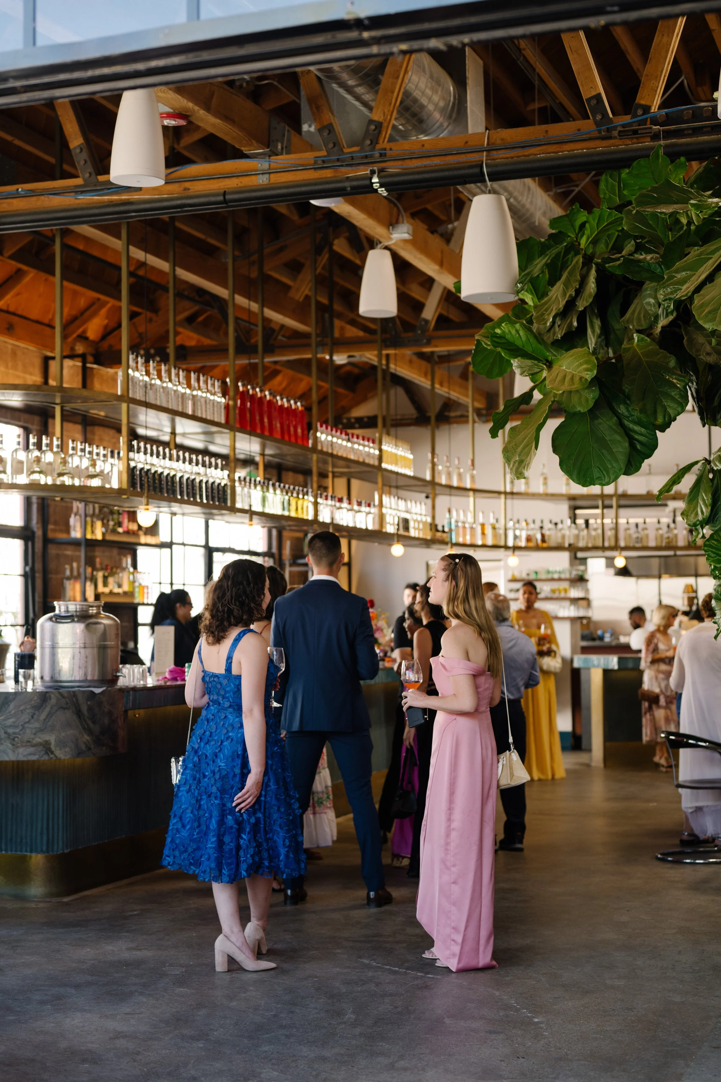People socializing at a bar inside a stylish wedding venue with a high wooden ceiling, hanging white lamps, and a large green plant.
