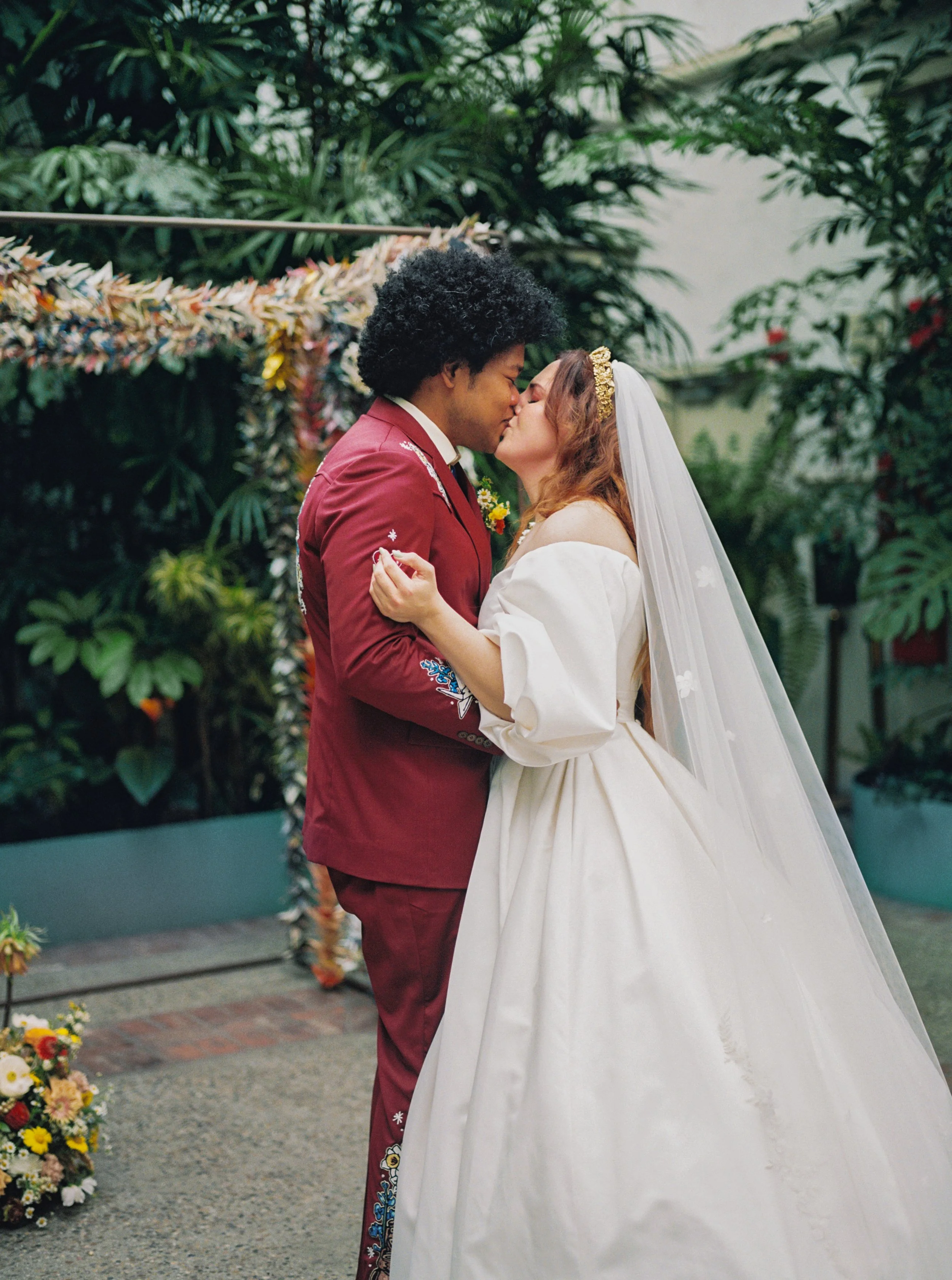 A newlywed couple sharing a kiss inside a lush, green conservatory or botanical garden, with floral decorations in the background.
