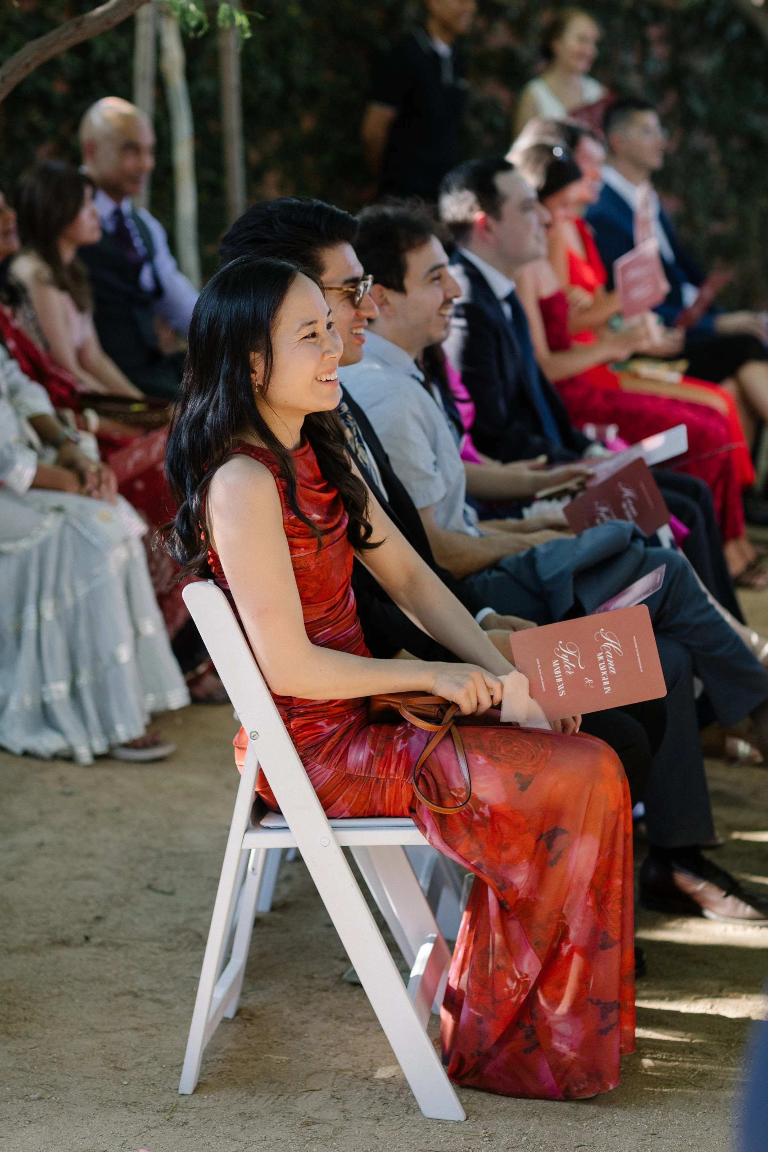 A woman in a red dress sitting on a white folding chair, smiling, at an outdoor wedding ceremony with other seated attendees.