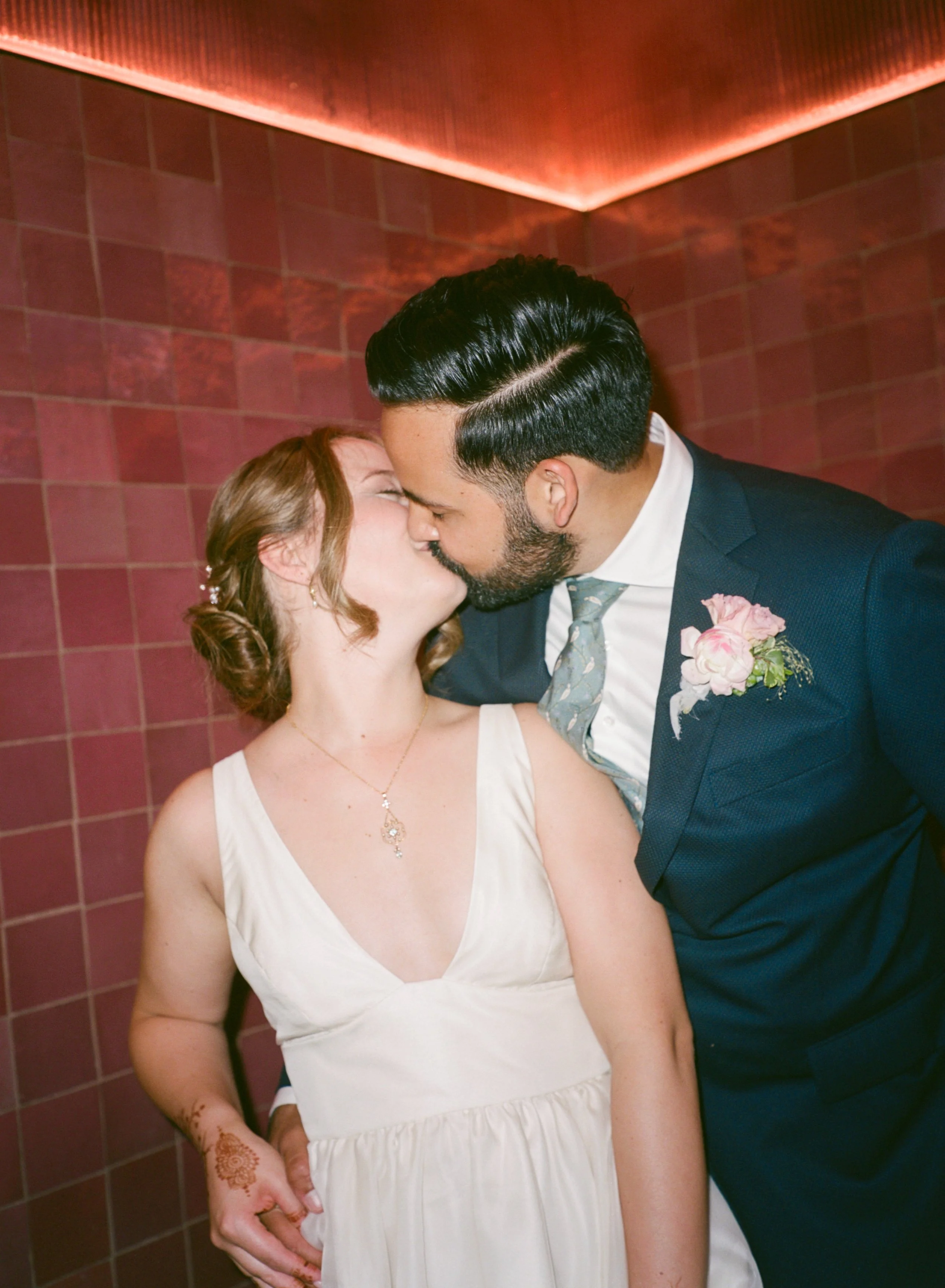 A couple is kissing at their wedding, the woman wears a white dress with henna on her hand and a necklace, the man in a dark suit with a boutonniere, against a pink tiled background.