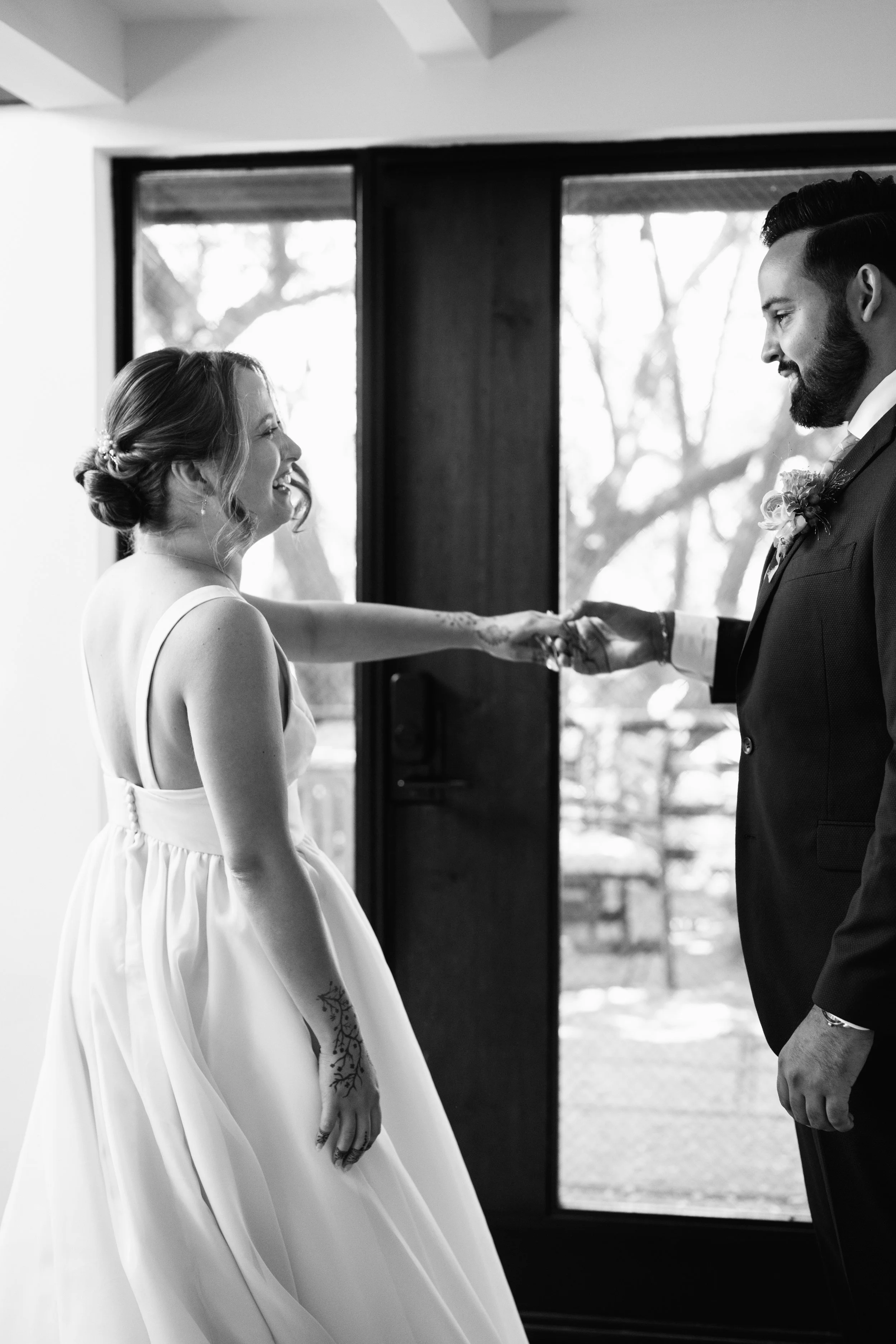 A bride and groom holding hands during the first look at their wedding, smiling at each other inside a room with large windows.