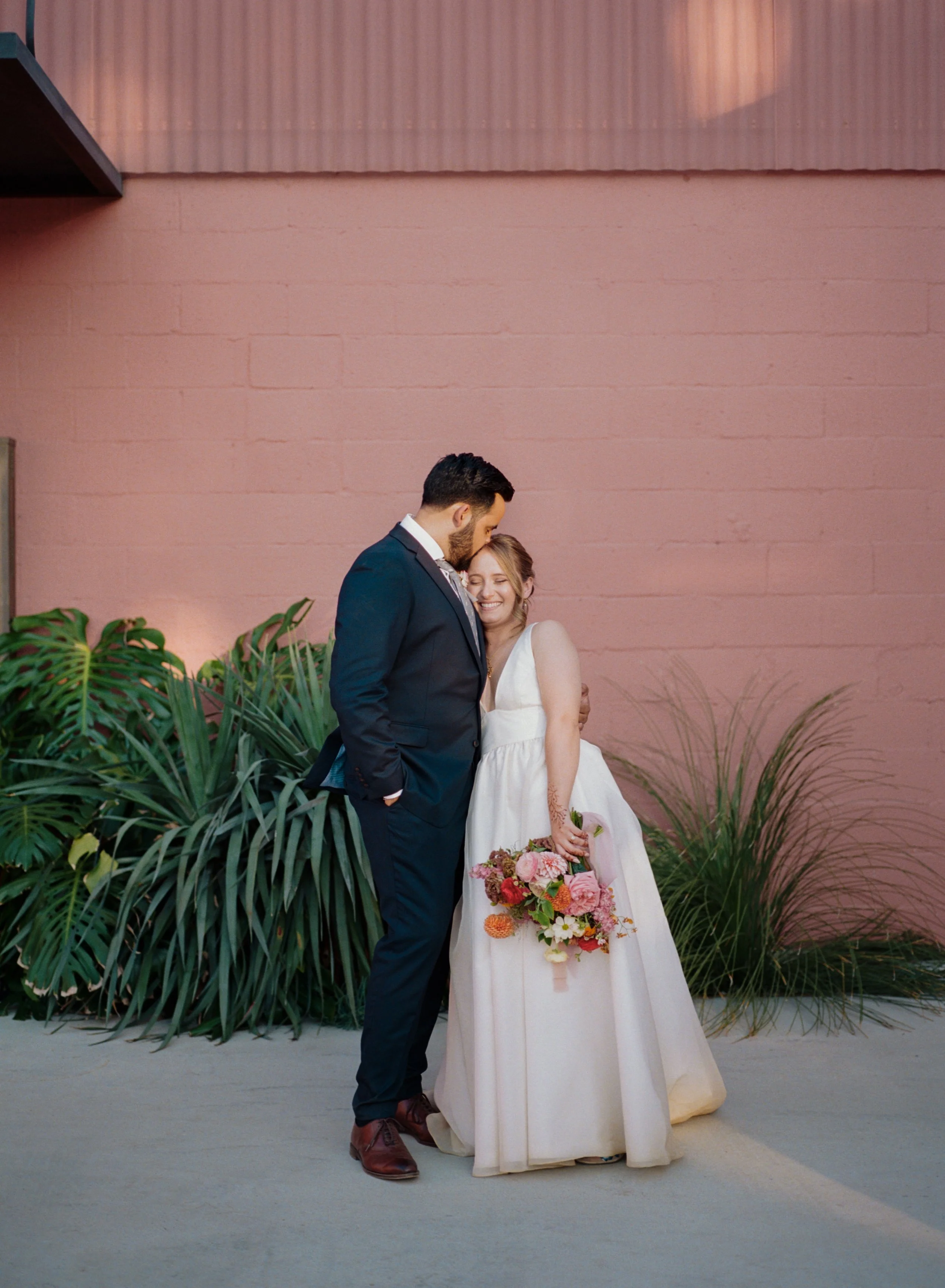 A bride and groom stand close together, smiling, with the groom's head resting on the bride's forehead. The bride wears a white wedding dress and holds a colorful bouquet of flowers. They stand outdoors against a pink painted wall with lush greenery