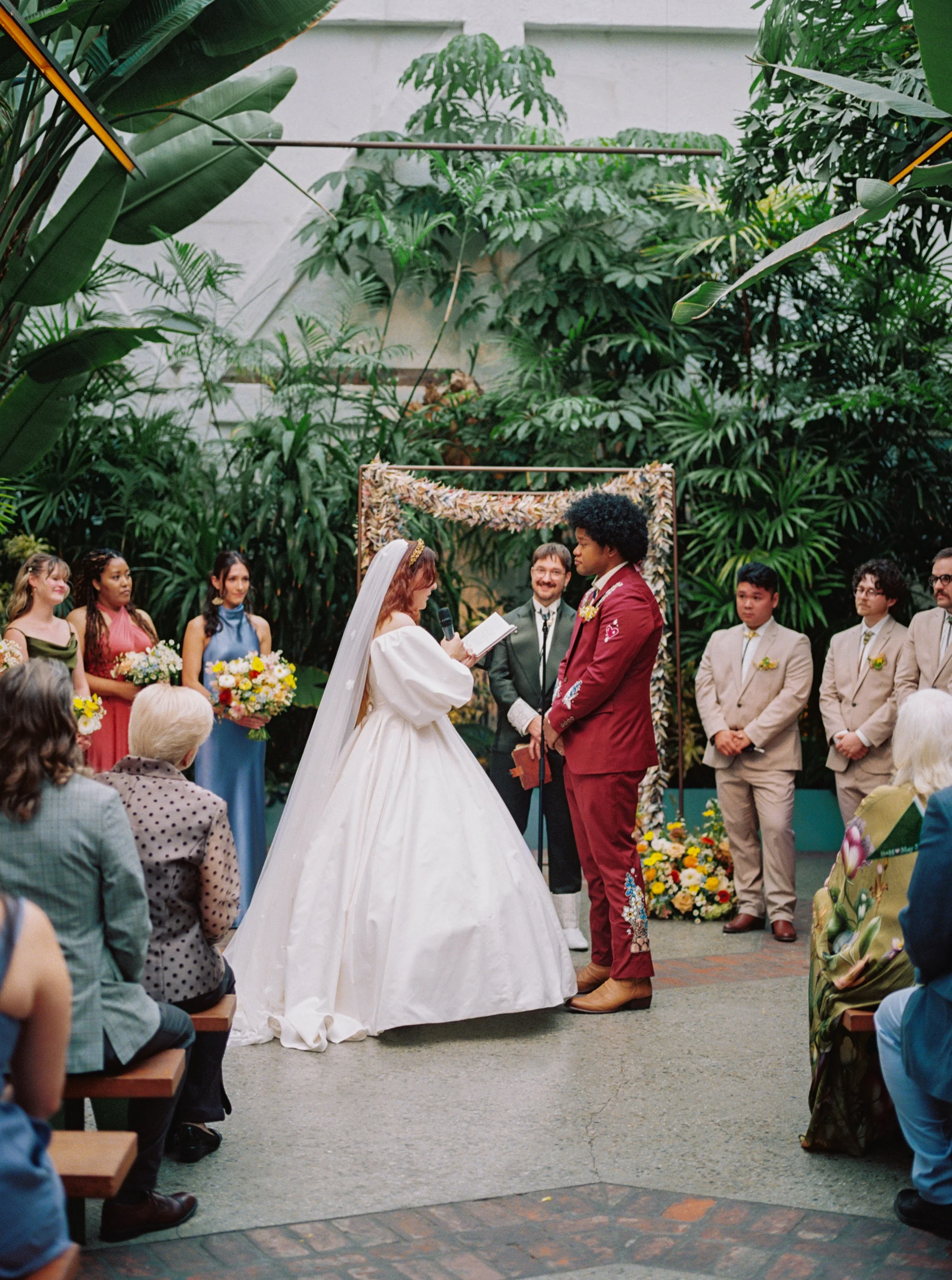 A wedding ceremony taking place in a lush indoor garden with tropical plants, featuring a bride and groom exchanging vows, surrounded by bridesmaids and groomsmen, with guests seated.