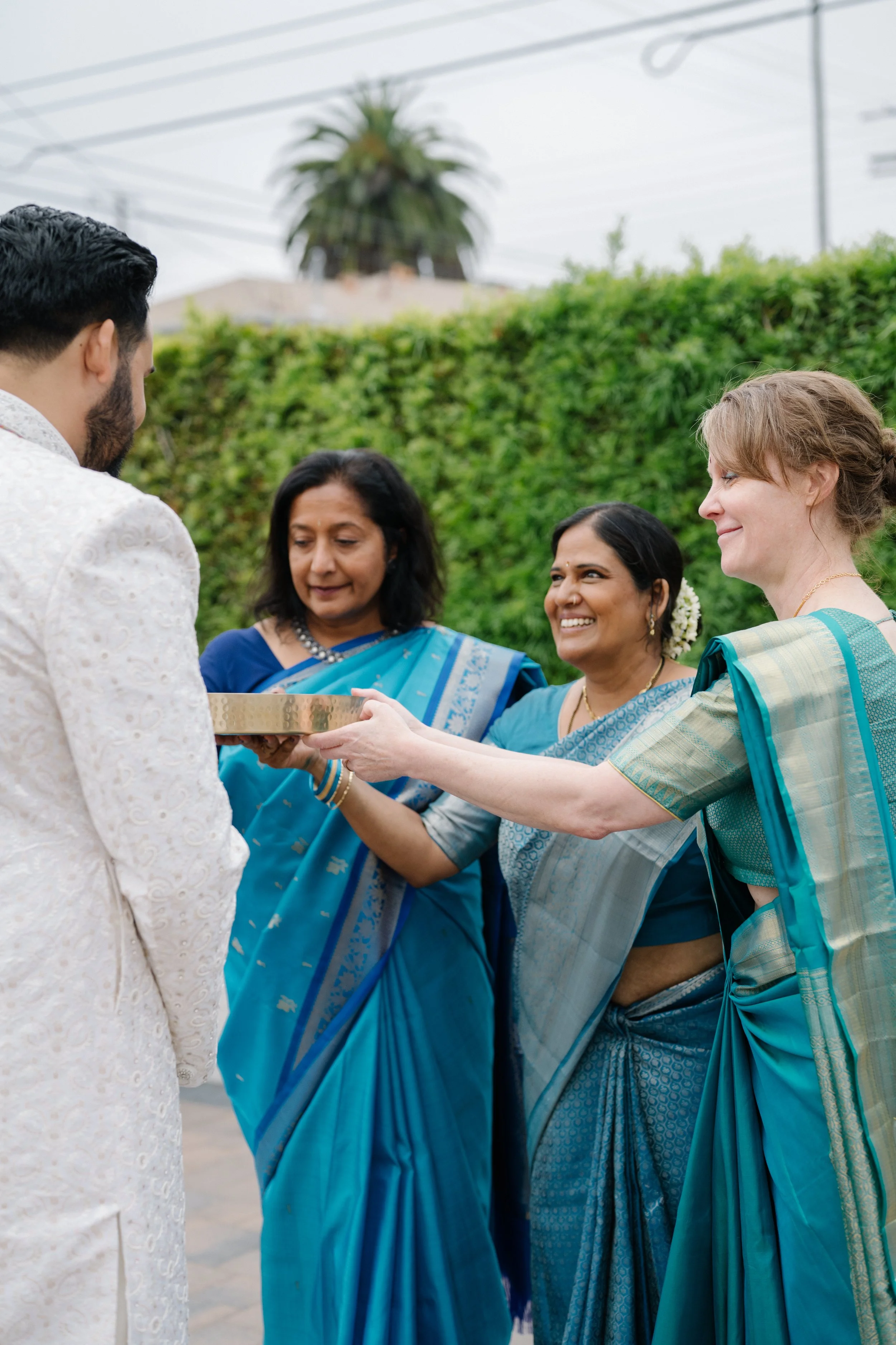 A traditional Indian wedding ceremony outdoors with a groom in a white sherwani and three women in blue and teal sarees, one holding a silver tray for a ritual during the baraat.