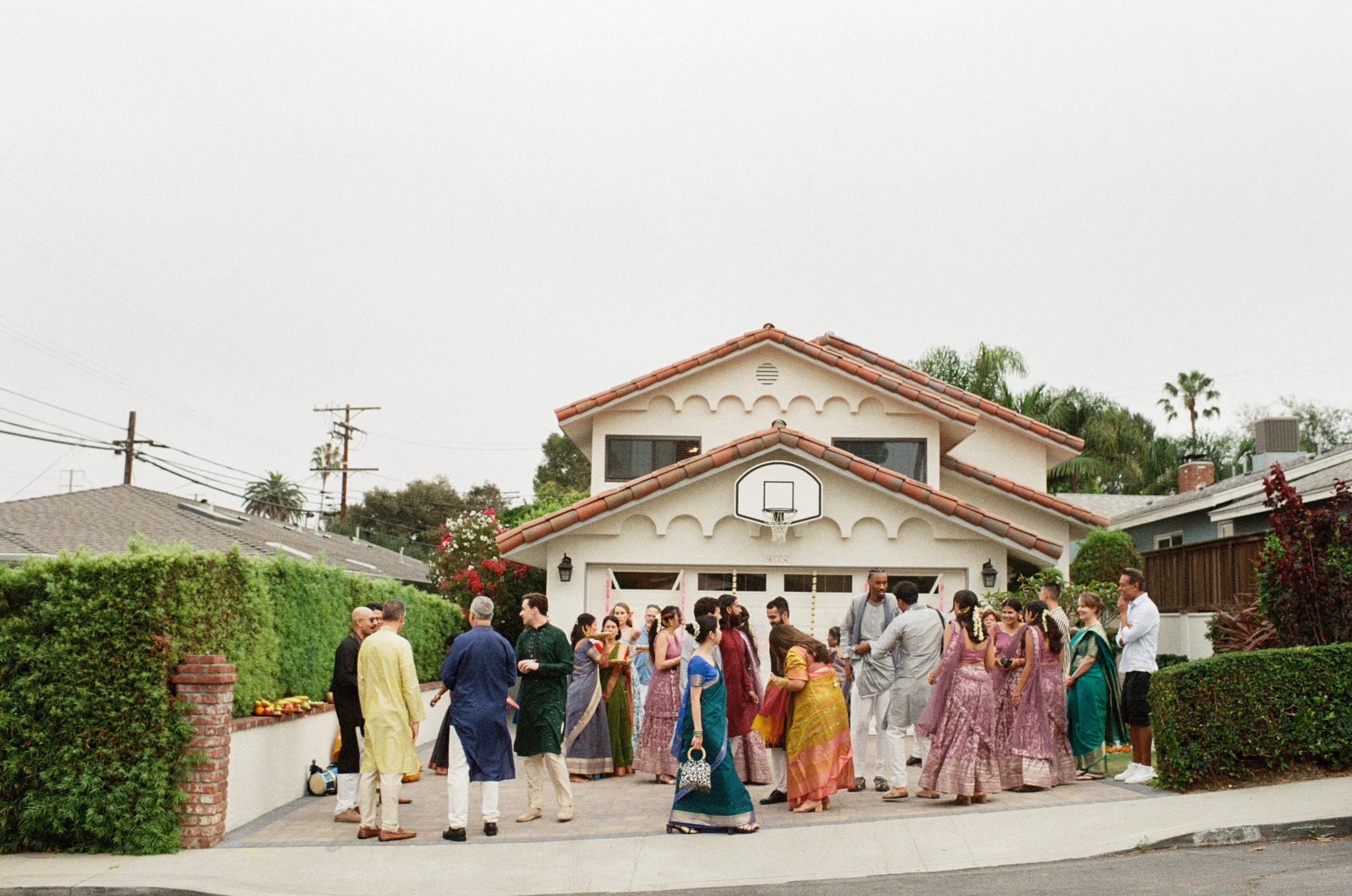 Group of people gathered outside a residential house, participating in an Indian baraat, with traditional Indian attire and food on a table.
