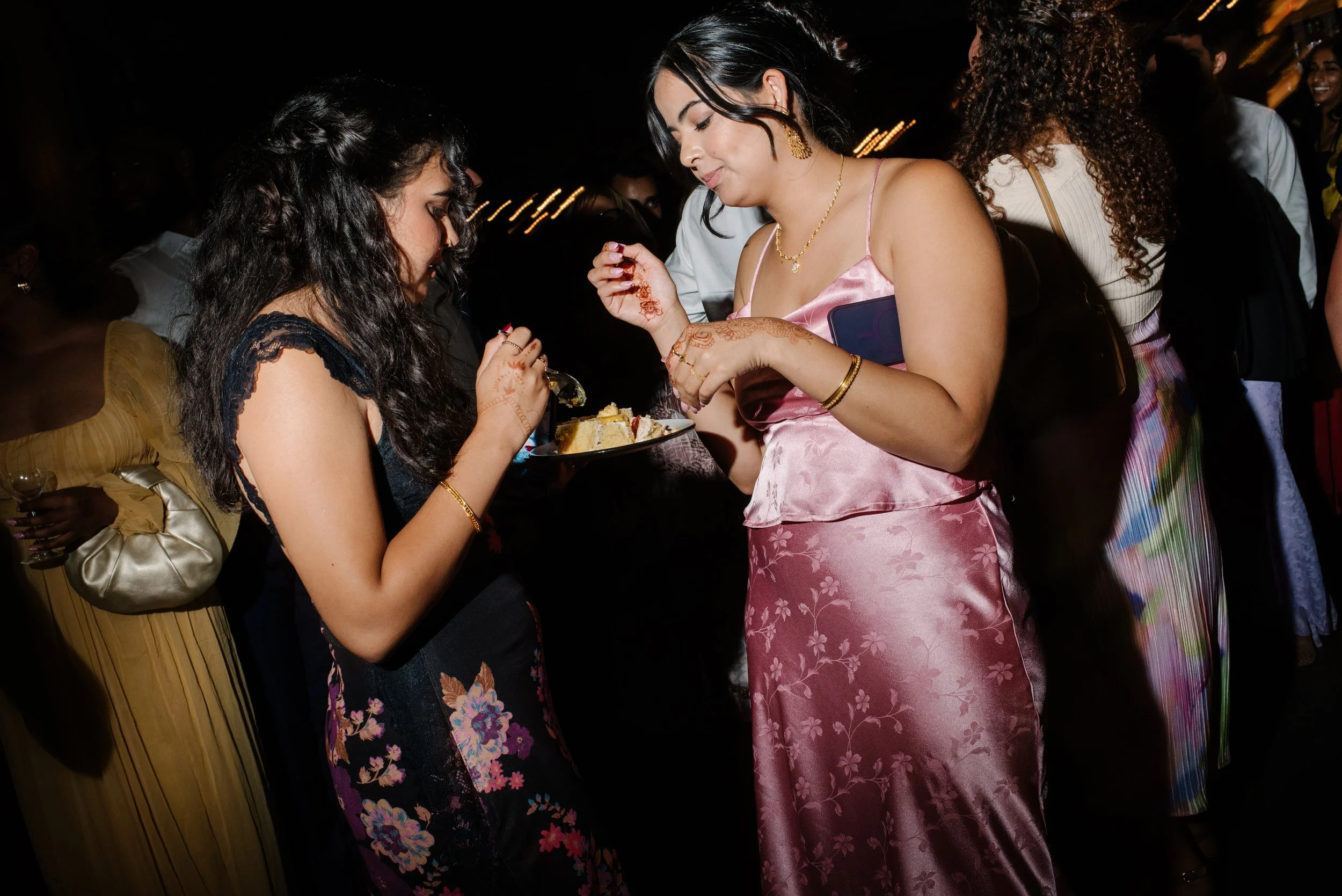 Two women at a celebration, one in a black floral dress and the other in a pink satin dress, sharing cake, surrounded by guests in party attire, in a dimly lit setting.