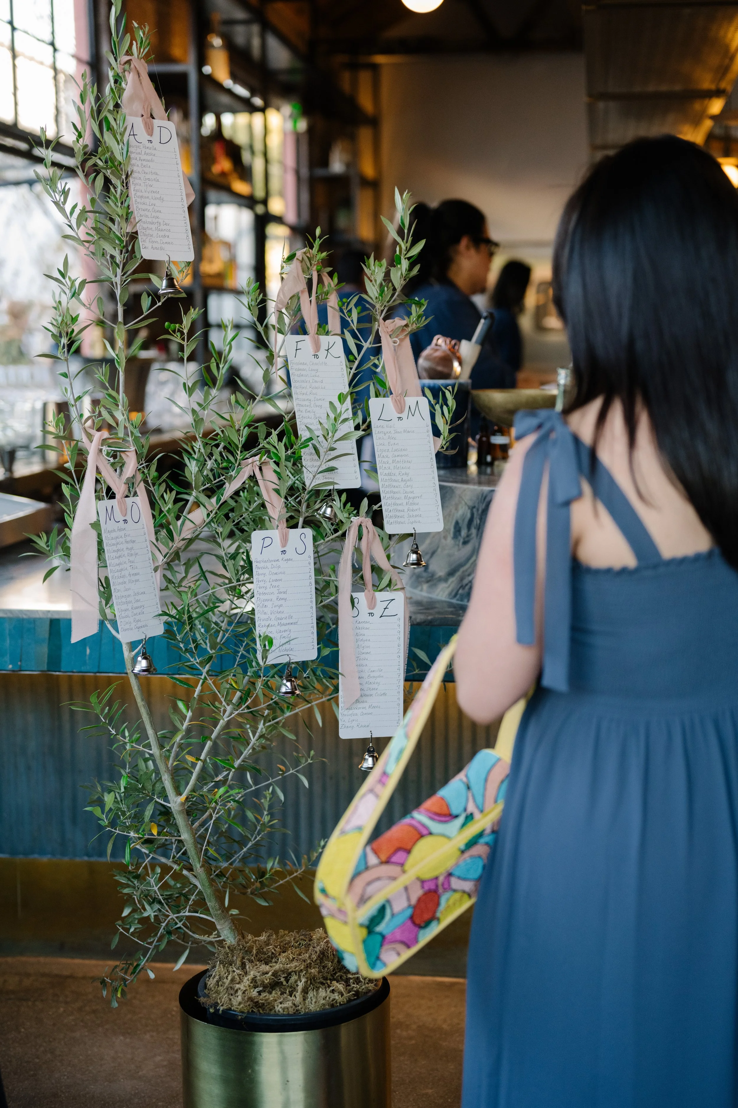 A woman in a blue dress stands in front of a decorated olive tree with hanging table assignments for a wedding dinner.