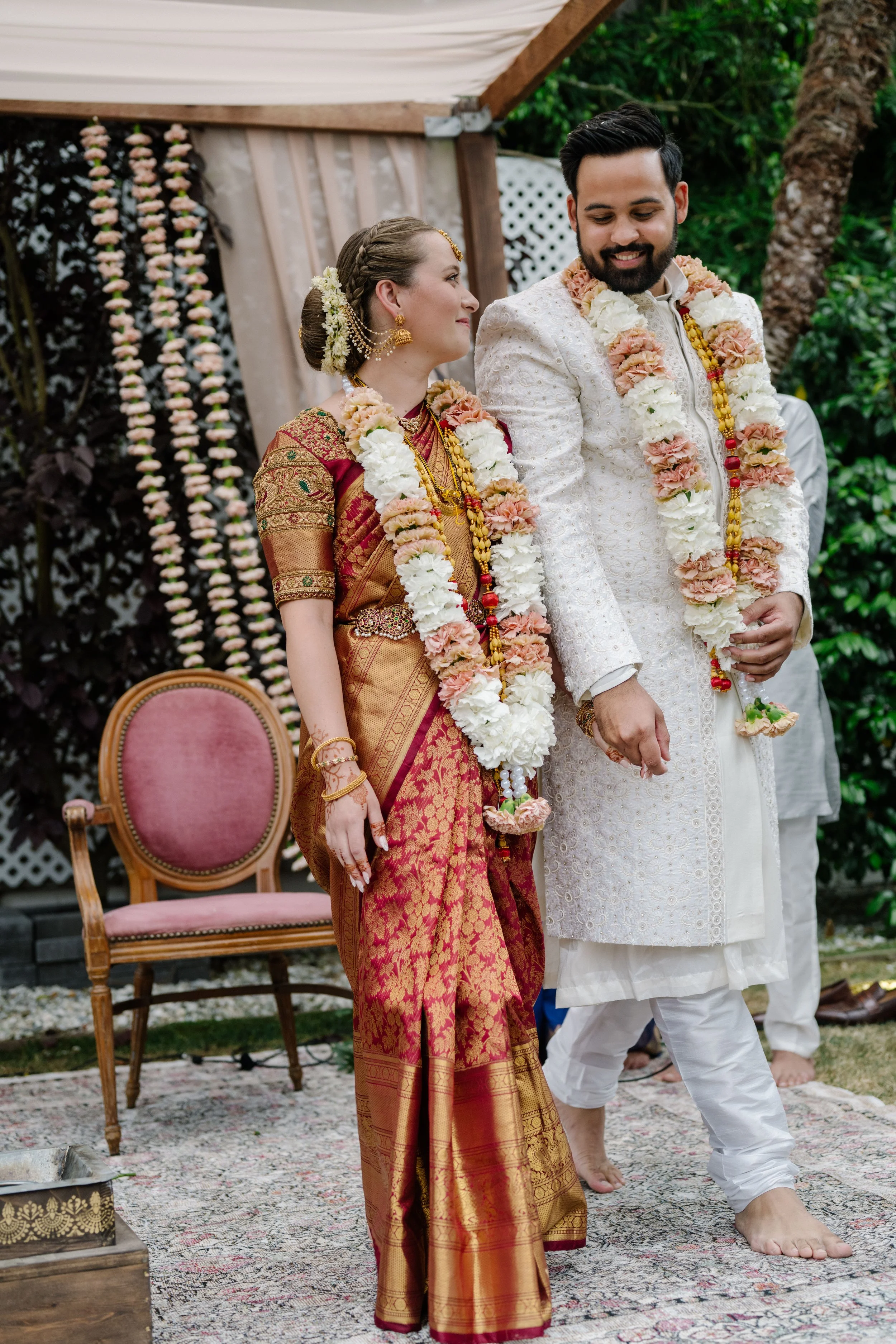 A couple in traditional Indian wedding attire celebrating outdoors, with the woman in a red and gold saree and the man in white embroidered kurta, both wearing flower garlands.