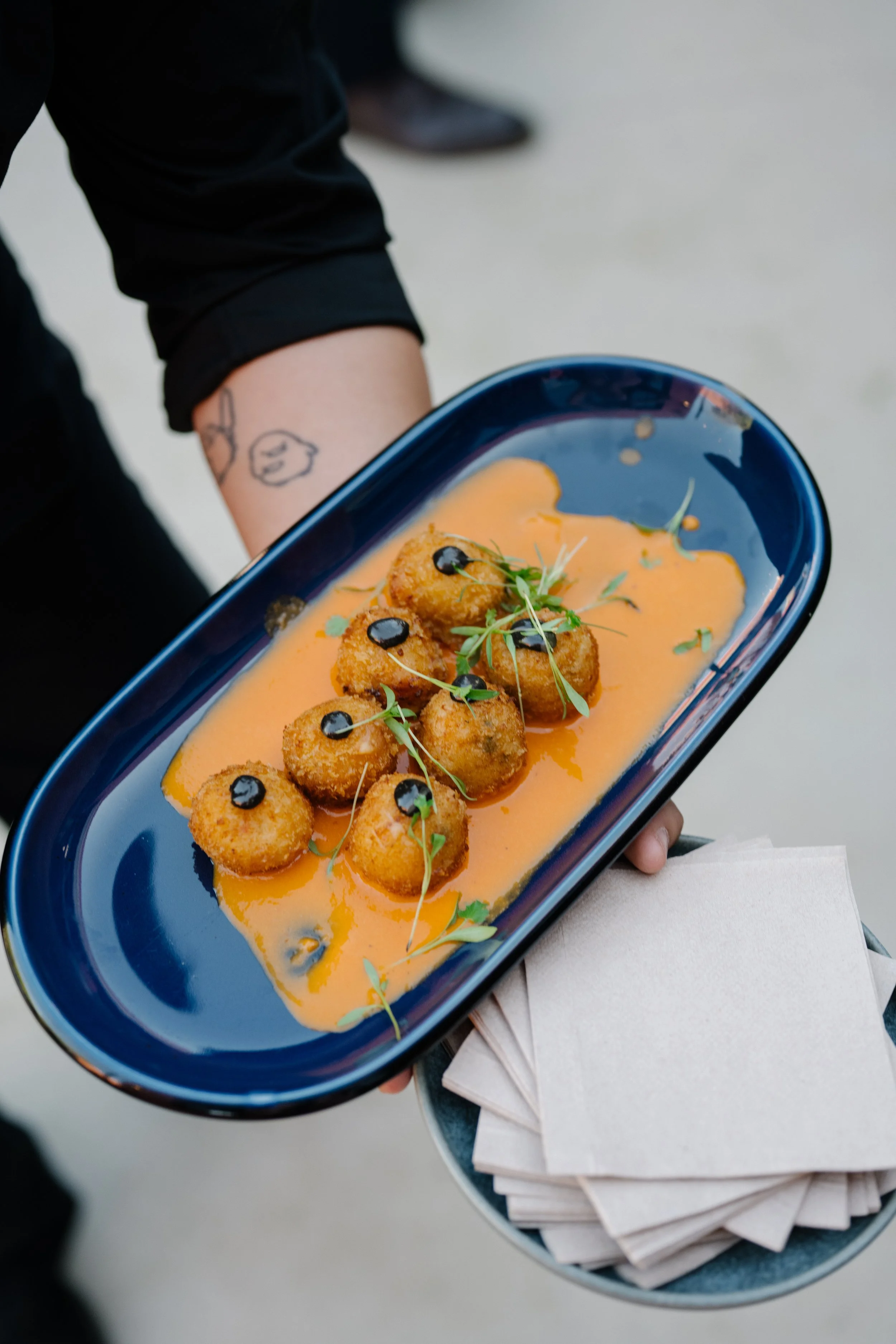 Person holding a blue dish with breaded appetizers drizzled with black sauce, garnished with microgreens on a bed of orange sauce, alongside white napkins on a small gray tray.