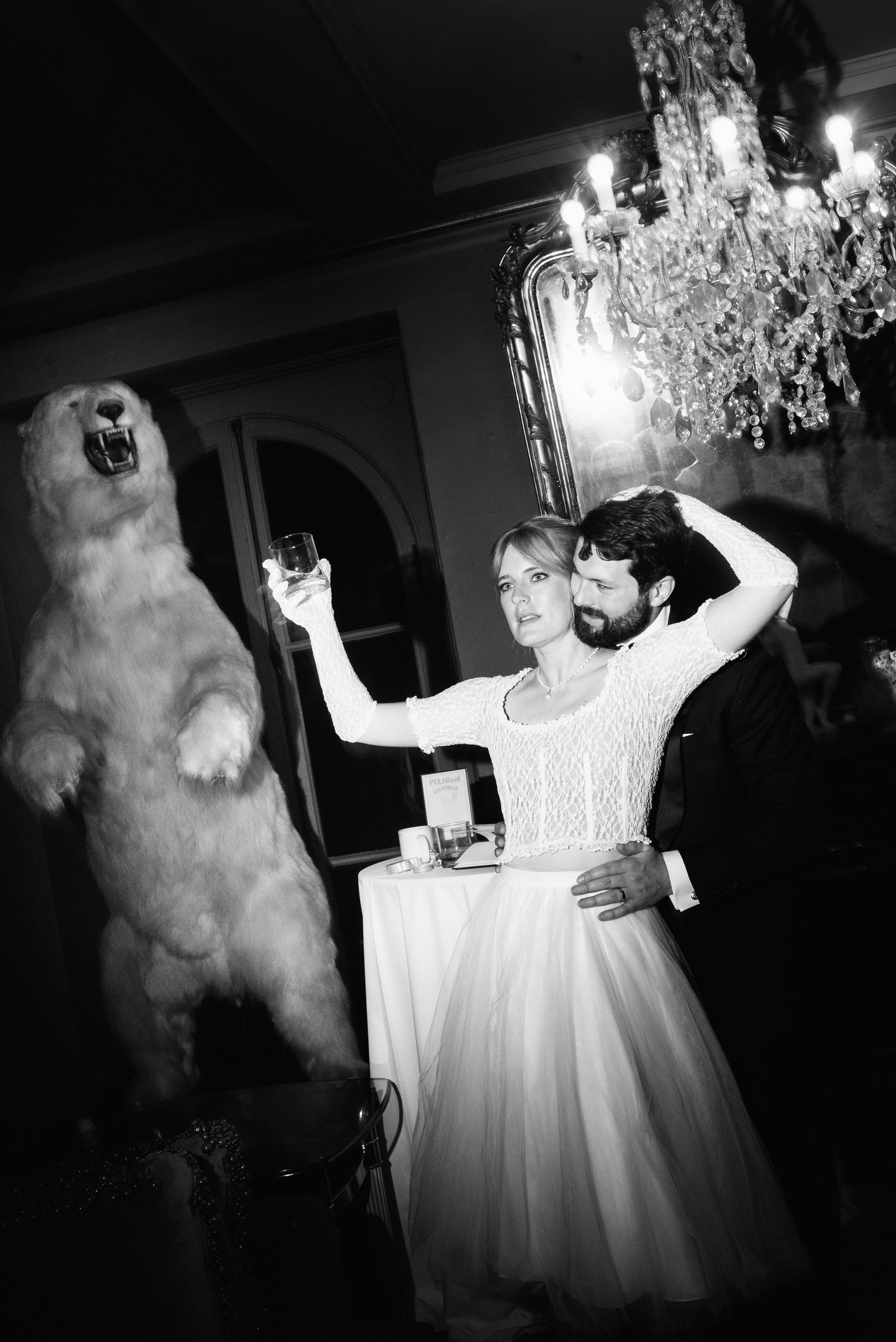 A woman in a wedding dress holding up a glass, while a man in a tuxedo wraps his arms around her waist from behind. A taxidermied polar bear stands nearby a chandelier and ornate mirror. The photo is black and white with heavy direct flash.