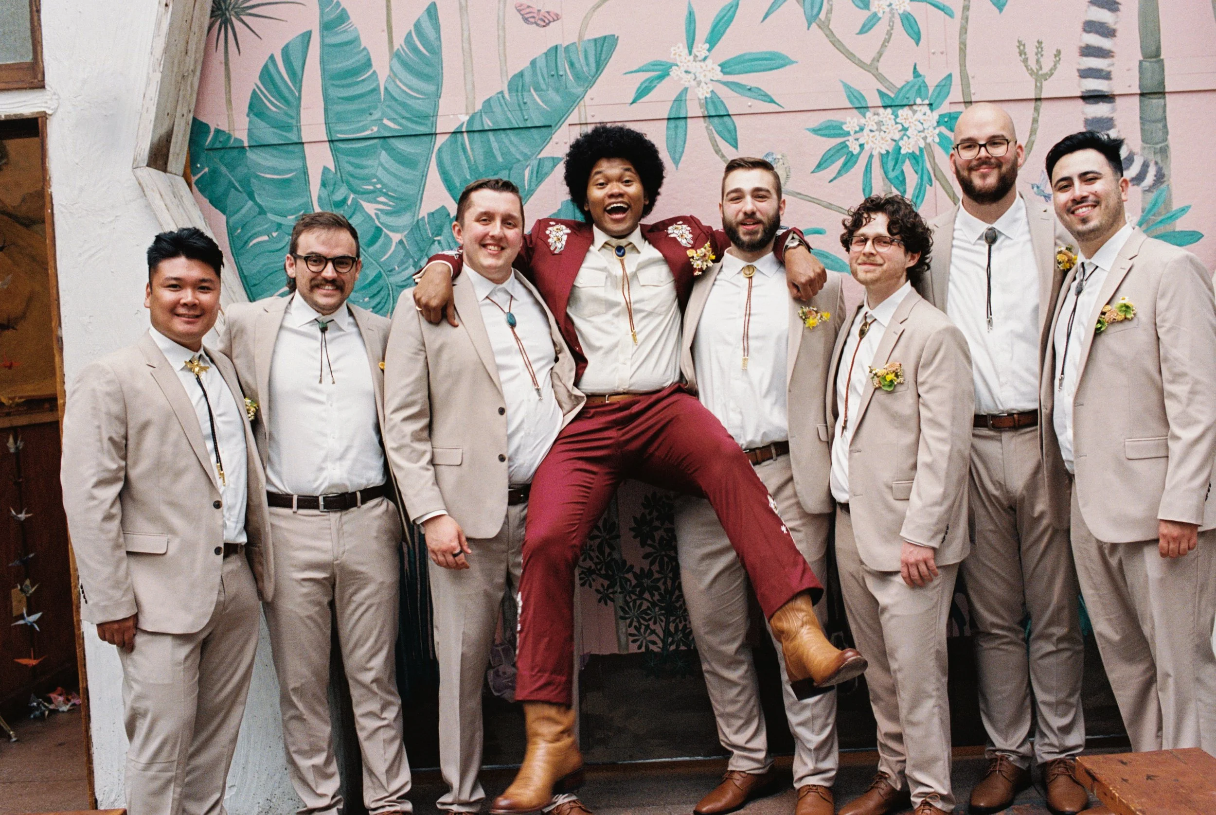 A group of nine men dressed in light-colored suits and white shirts, standing together and smiling, with groom wearing a red outfit and jumping with excitement. They are in front of a mural with large blue and green leaves and pink background.