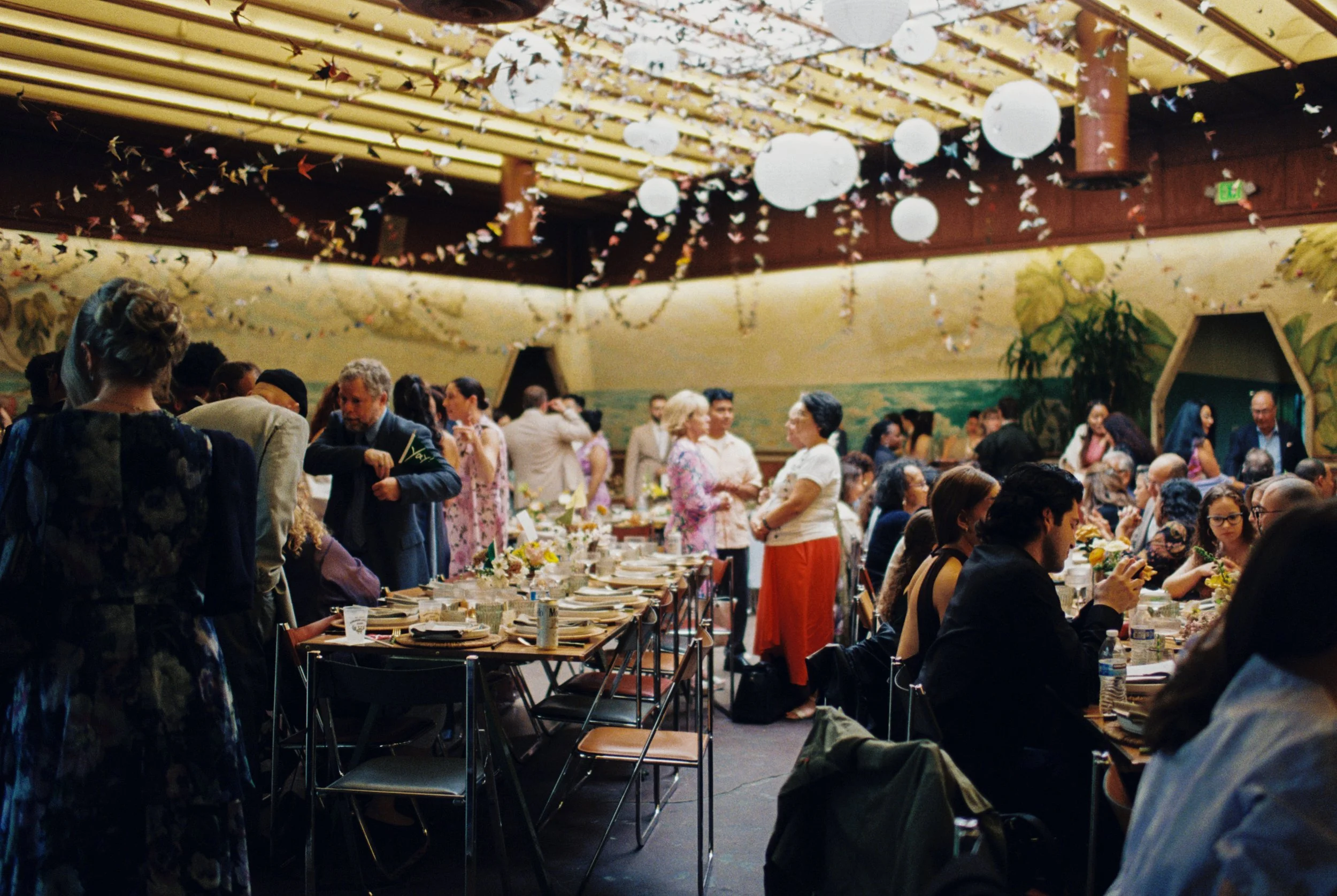 A crowded indoor wedding with people socializing around decorated tables, hanging paper lanterns, and origami cranes hanging from the ceiling.