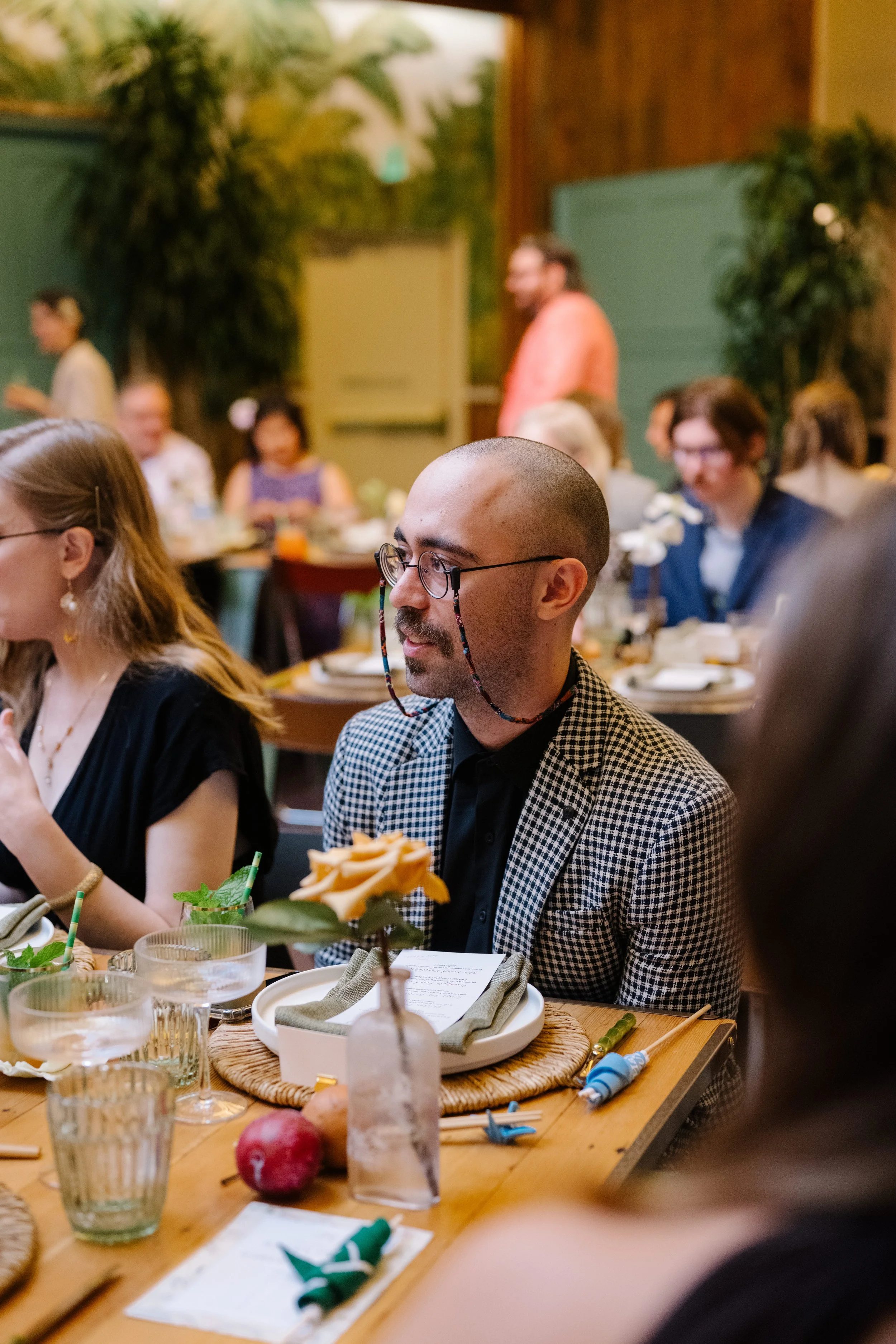 A man with glasses and a checked blazer sitting at a dinner table during a wedding, with other guests in the background, and a flower in a vase on the table.
