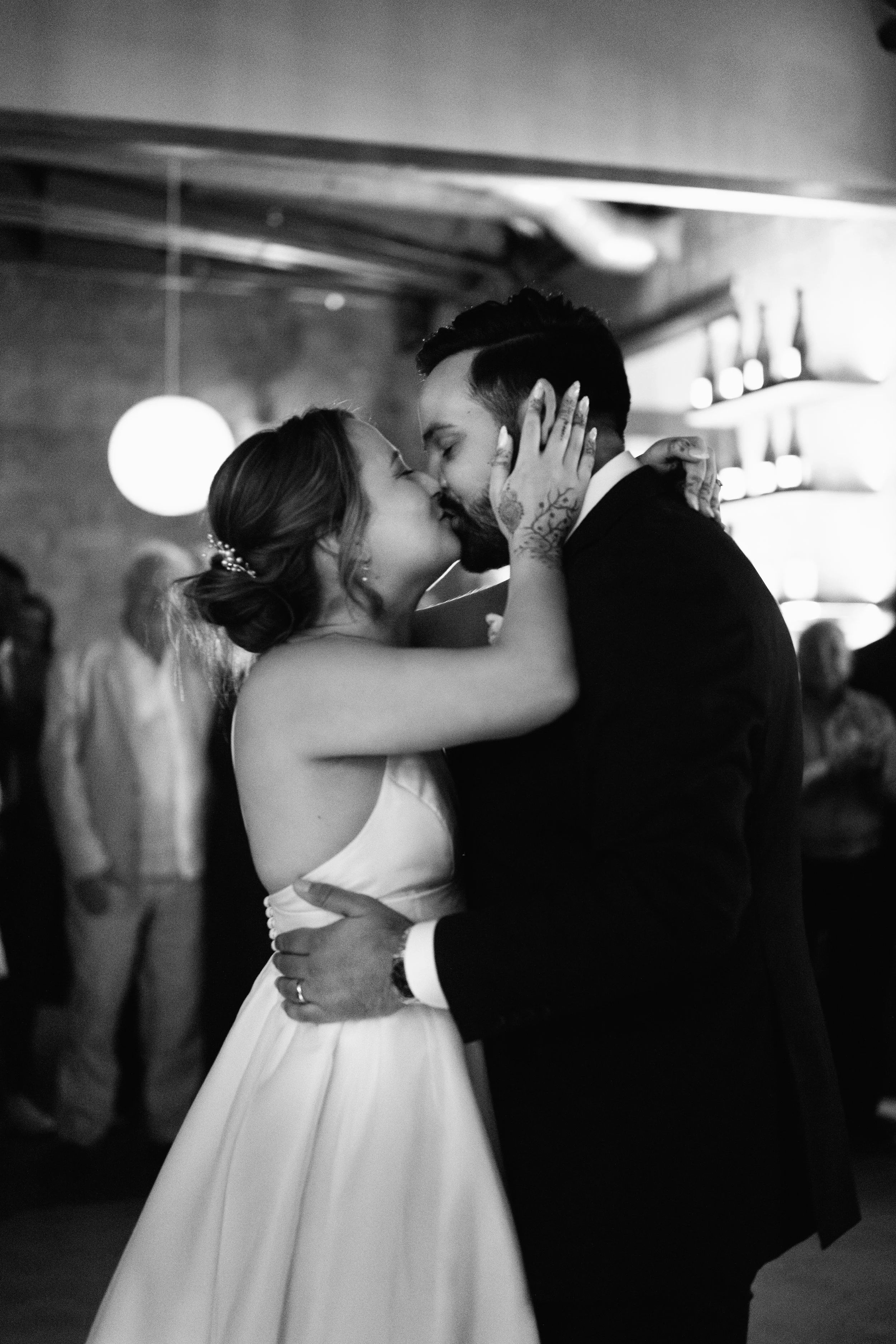 A black-and-white photo of a bride and groom sharing a kiss during their wedding reception, with guests in the background.